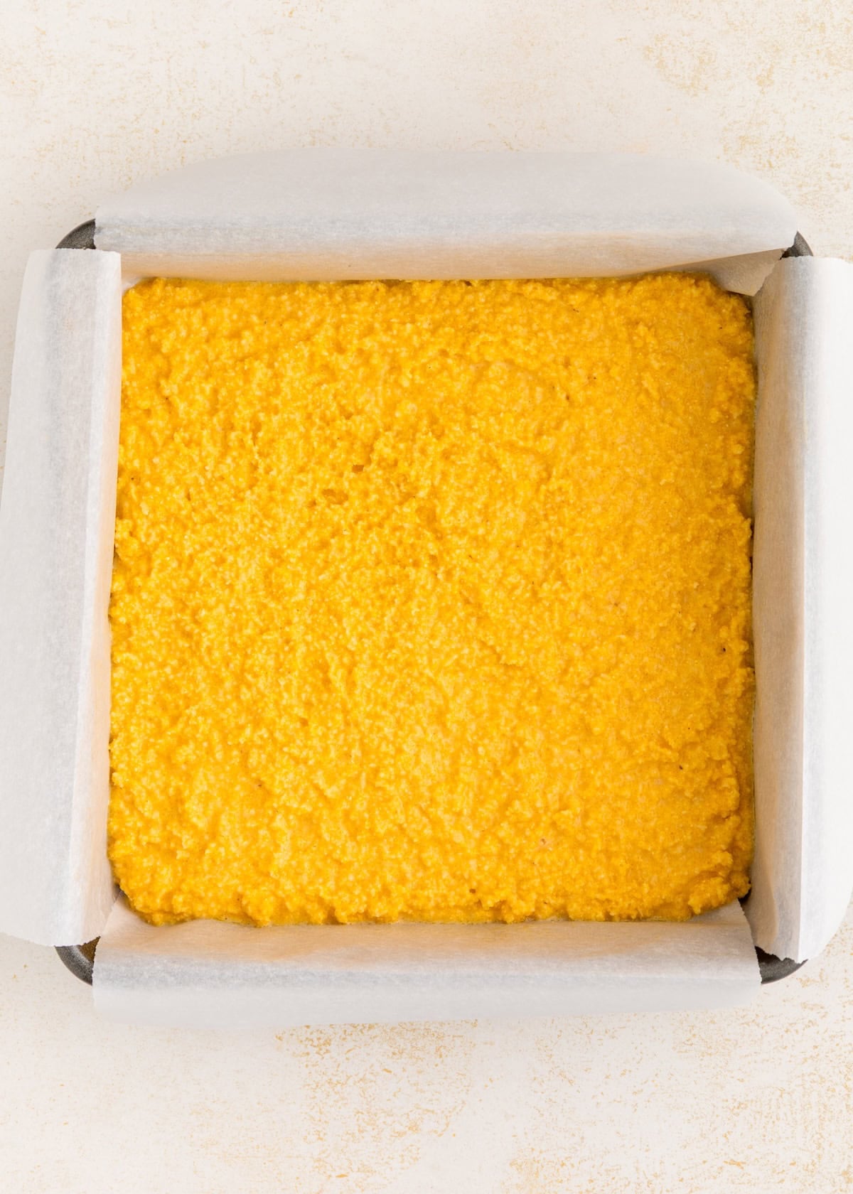 A square baking pan lined with parchment paper, filled with uncooked yellow cornmeal mixture.
