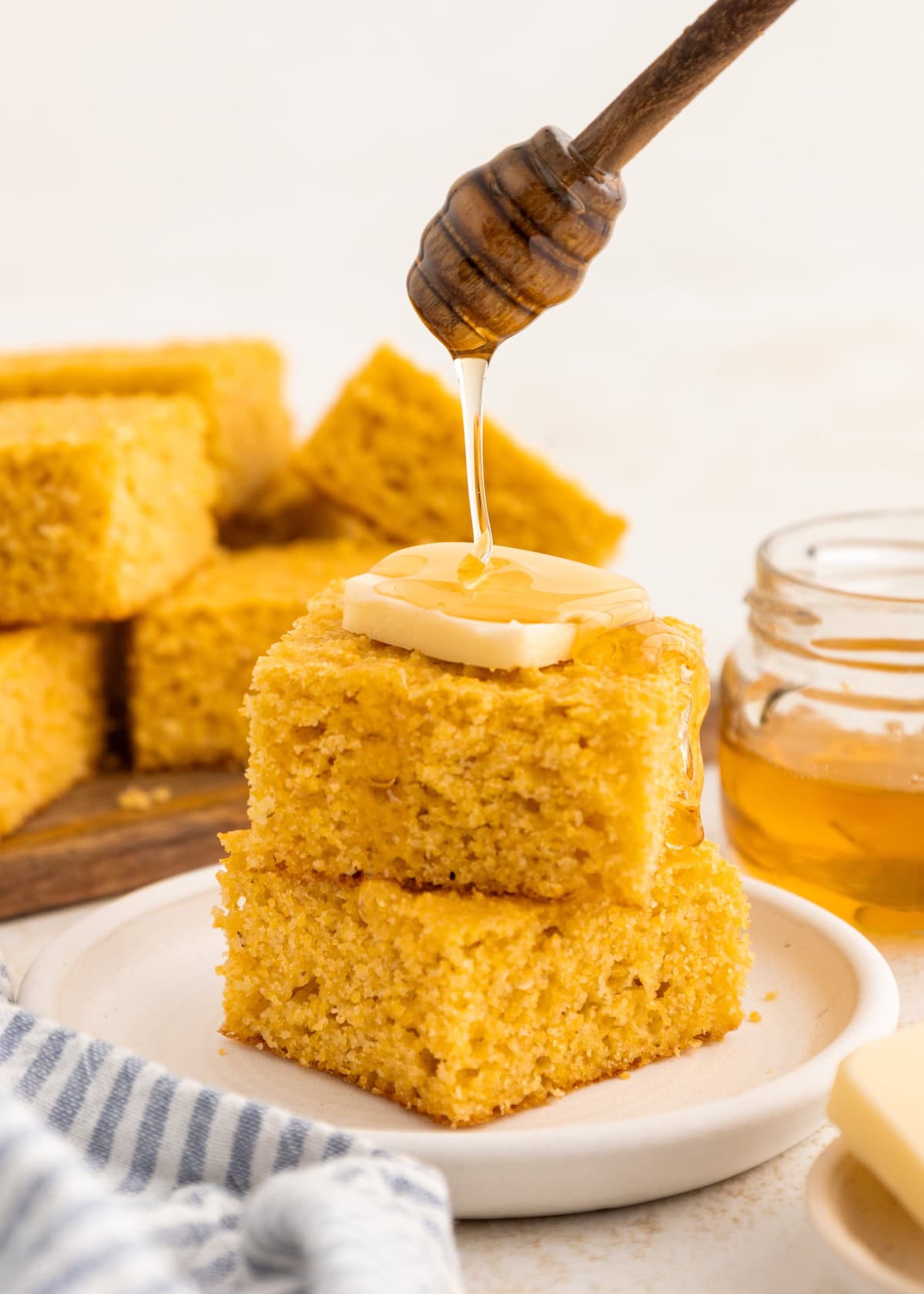 Two pieces of cornbread with butter, honey being drizzled on top, and a jar of honey nearby.