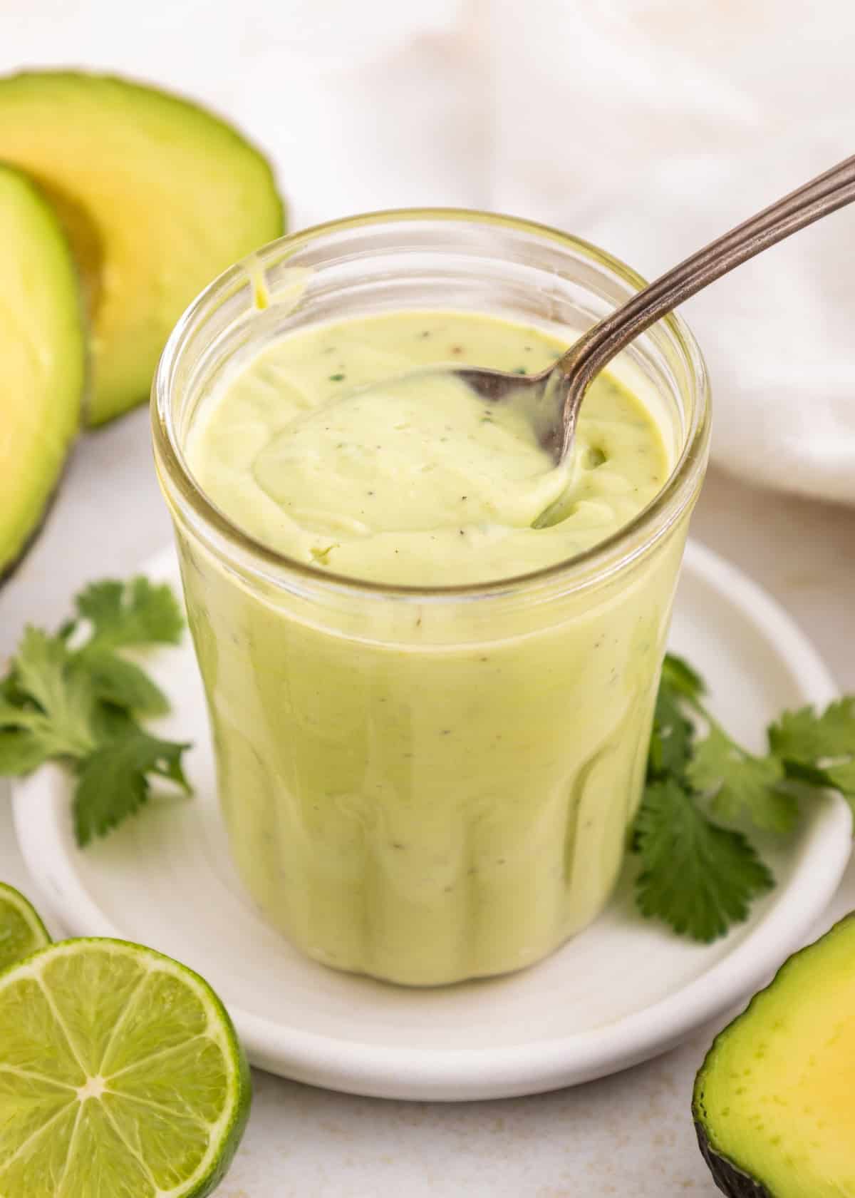 A jar of creamy avocado lime ranch dressing with a spoon, surrounded by lime and cilantro.