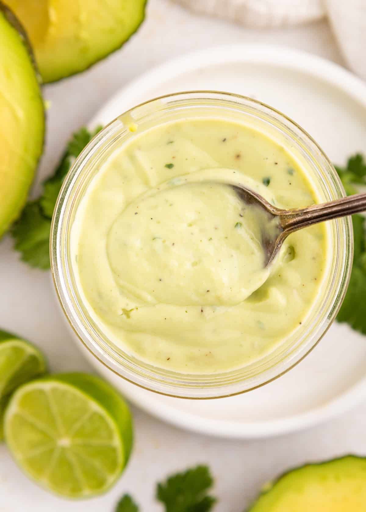 A spoon in a jar of creamy avocado lime ranch dressing, with lime and avocado halves nearby.