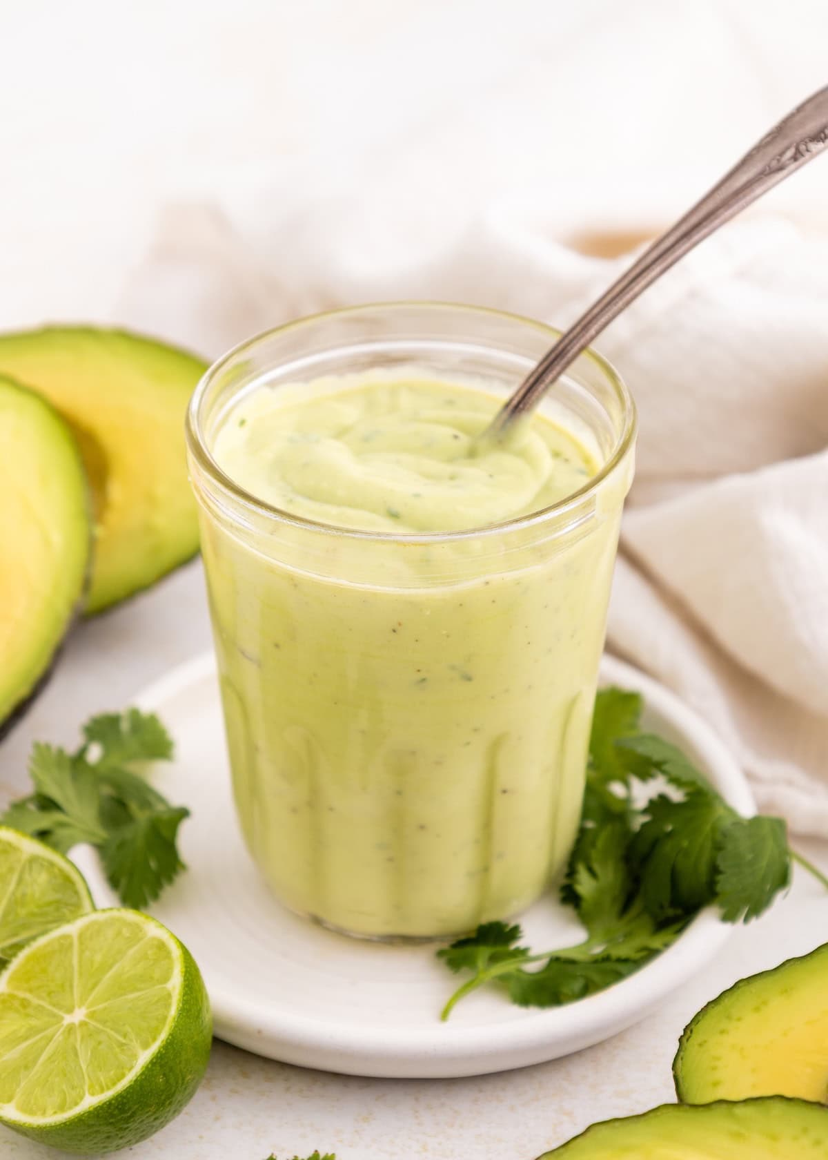 A glass of creamy avocado lime ranch dressing with a spoon, surrounded by avocado and cilantro.