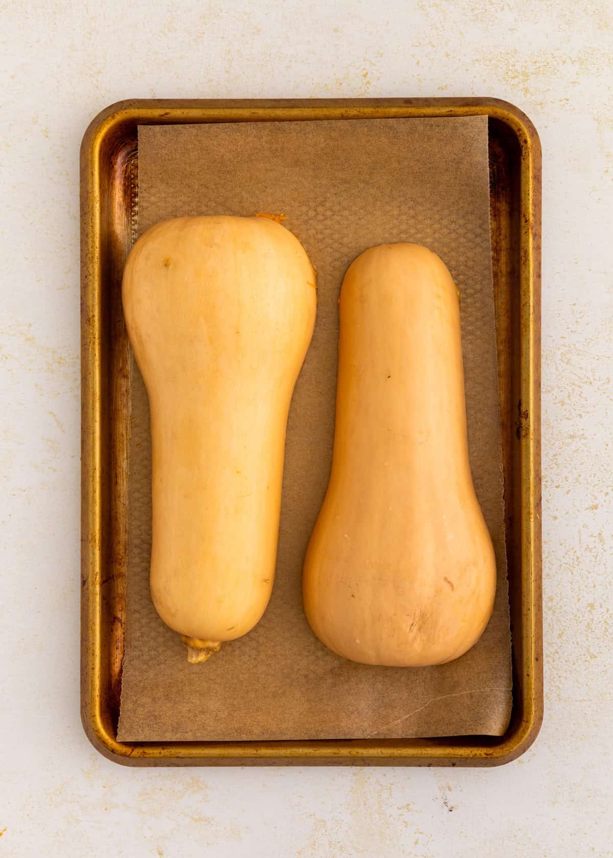 Two whole butternut squash on a parchment-lined baking sheet.