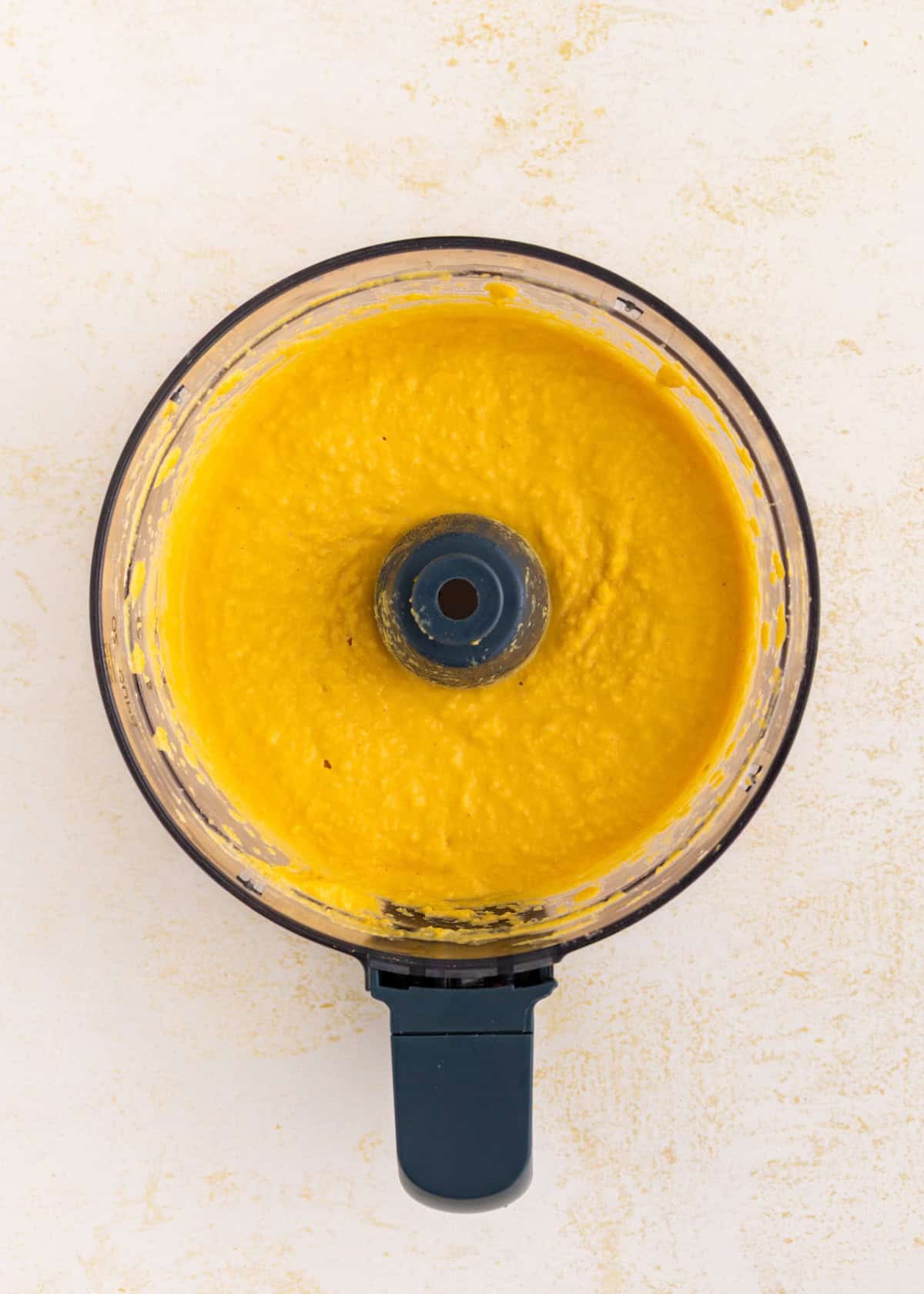 Top view of a food processor with bright yellow puree inside on a light surface.