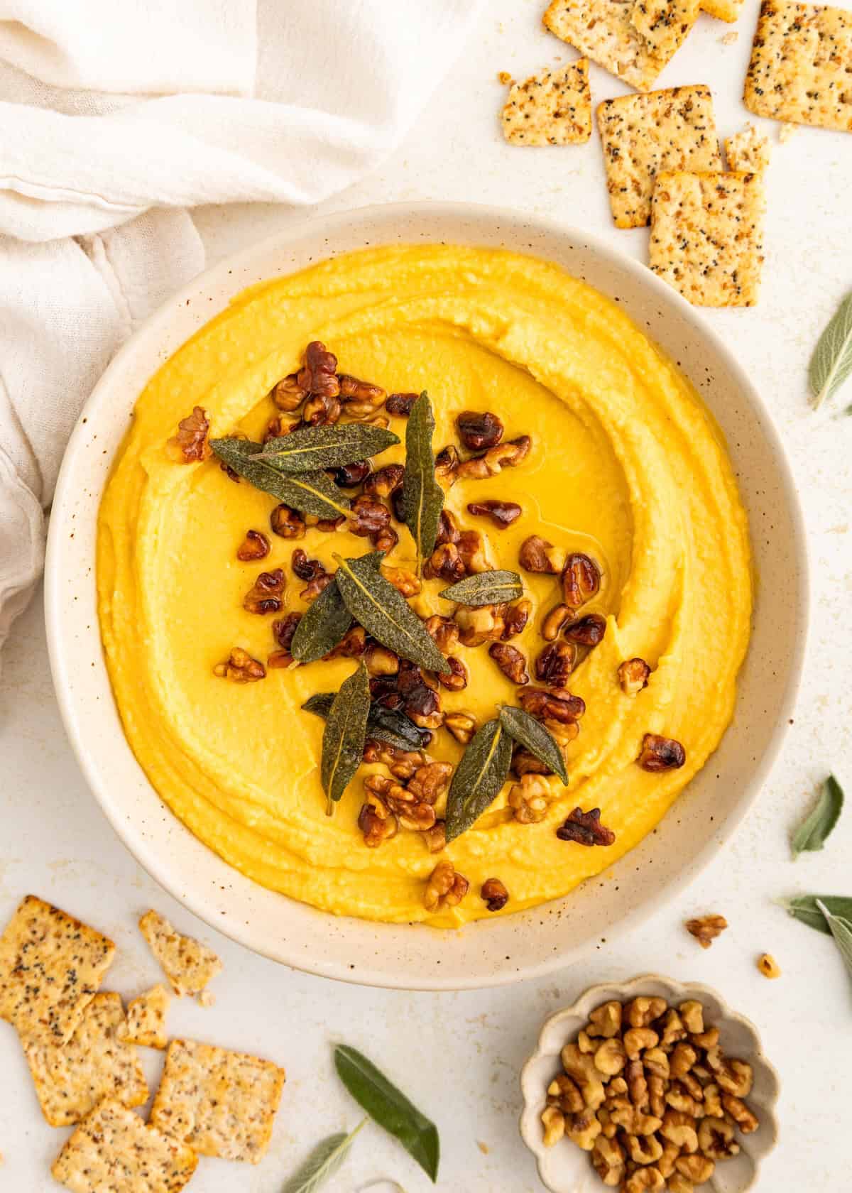 A bowl of yellow hummus topped with nuts and herbs, surrounded by crackers and sage leaves.
