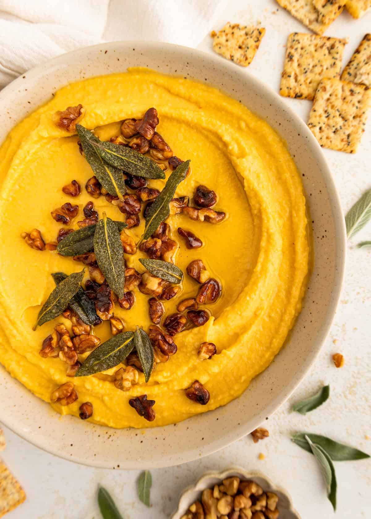 Creamy dip topped with walnuts and herbs in a bowl, surrounded by seeded crackers.