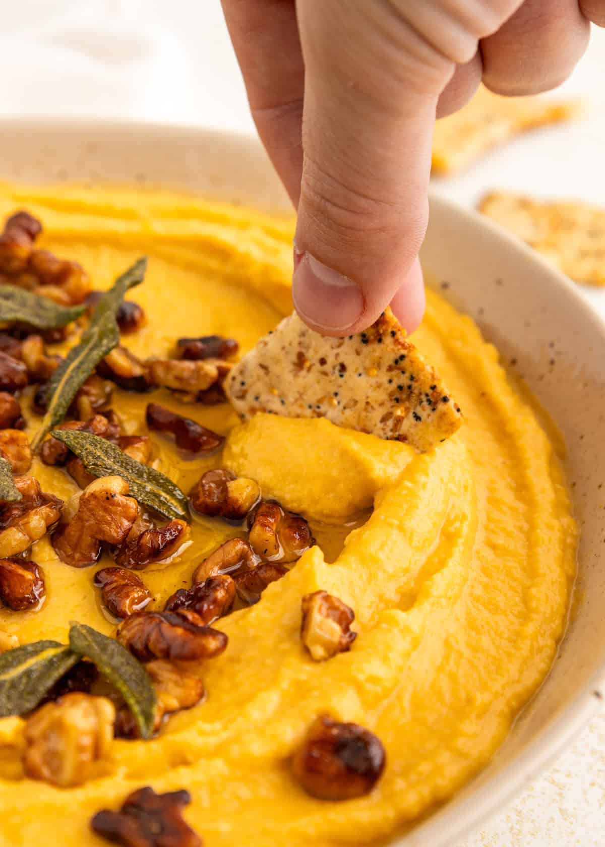A hand dips a cracker into creamy hummus topped with walnuts and herbs.