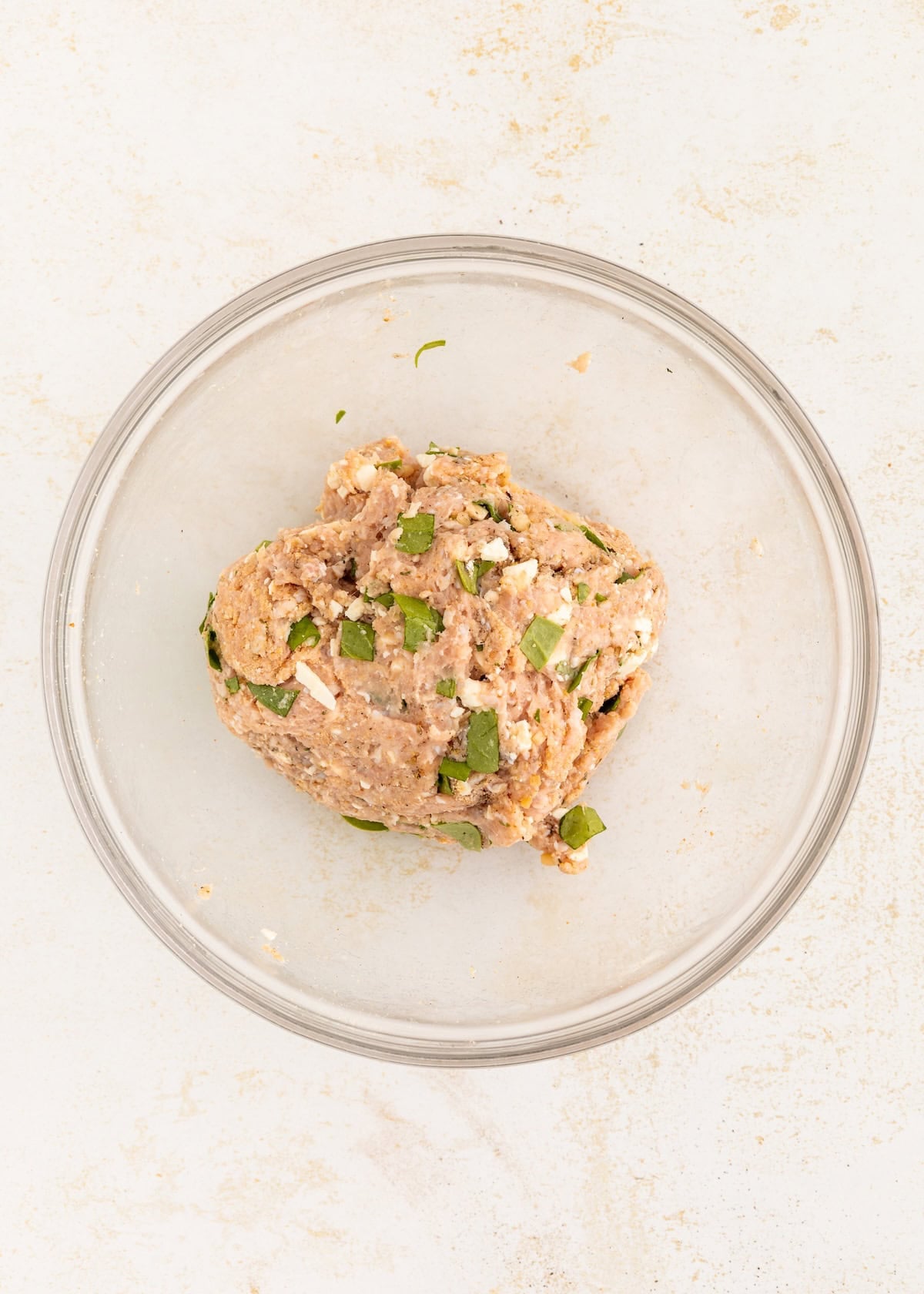 Ground meat mixture with herbs in a clear glass bowl on a light surface.