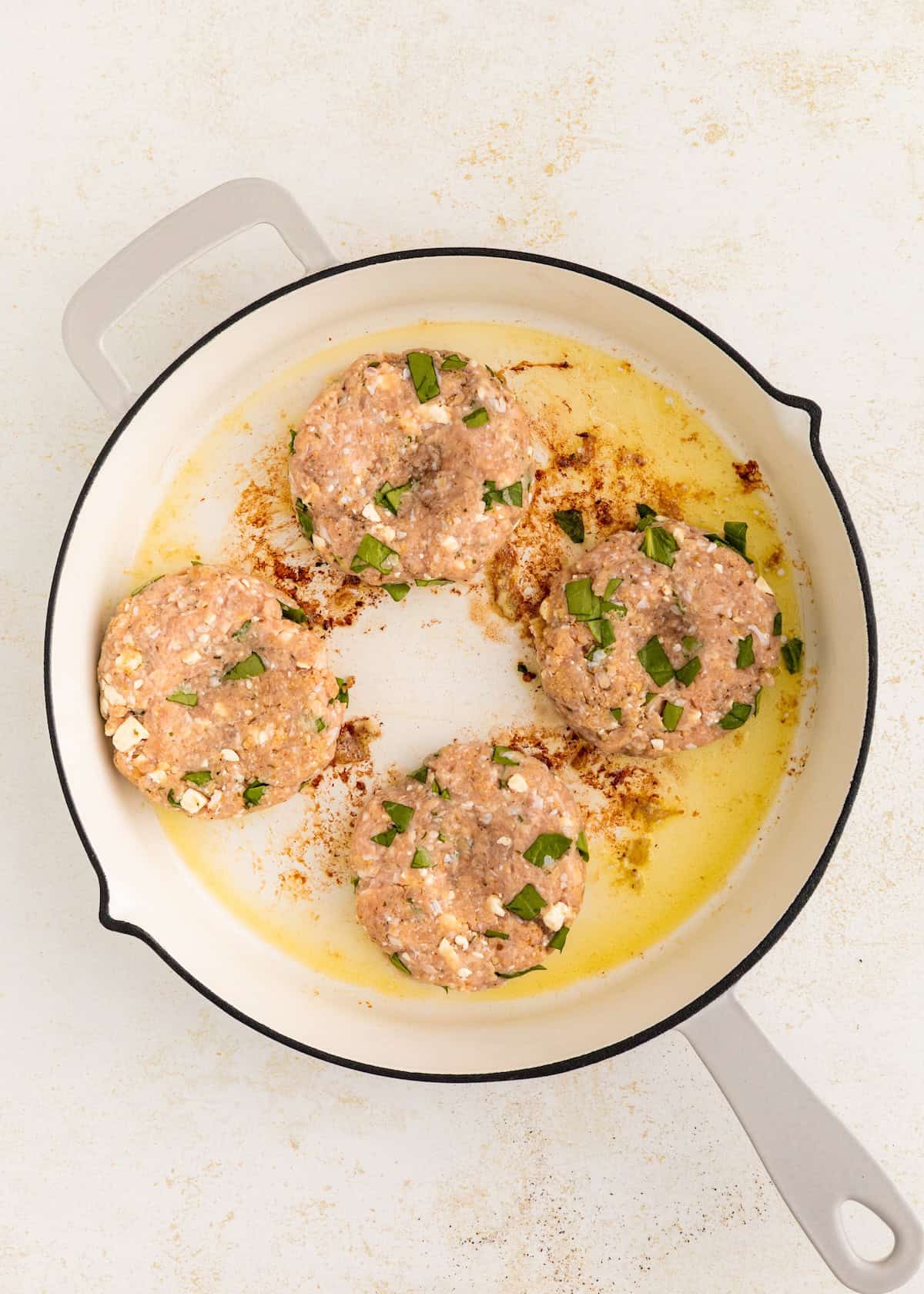 Four raw patties with green herbs are cooking in a white skillet with oil.