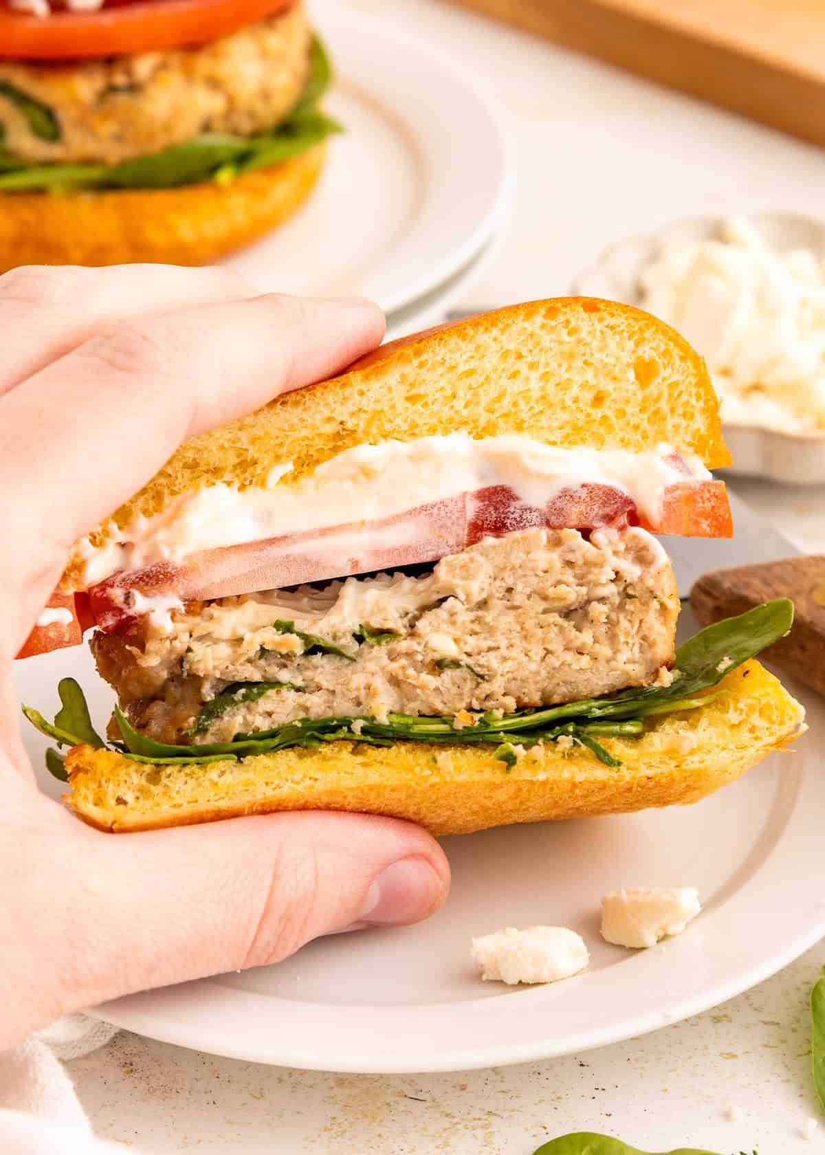 A hand holds a chicken burger with tomato, lettuce, and mayonnaise on a bun.