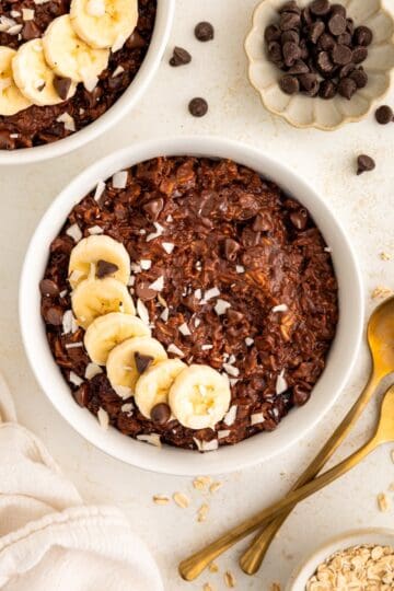 A bowl of chocolate oatmeal topped with banana slices and chocolate chips on a light surface.