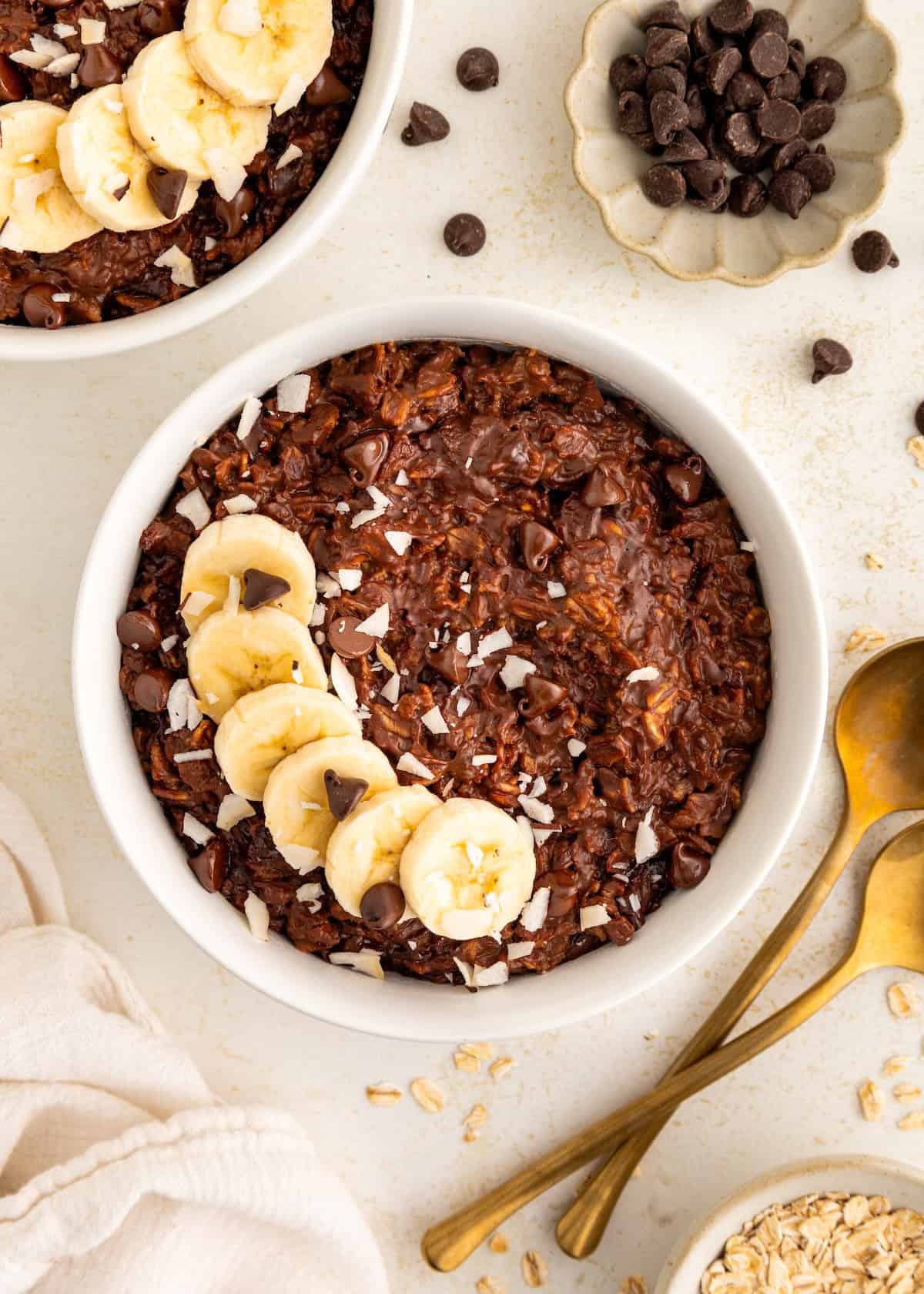 A bowl of chocolate oatmeal topped with banana slices and chocolate chips on a light surface.
