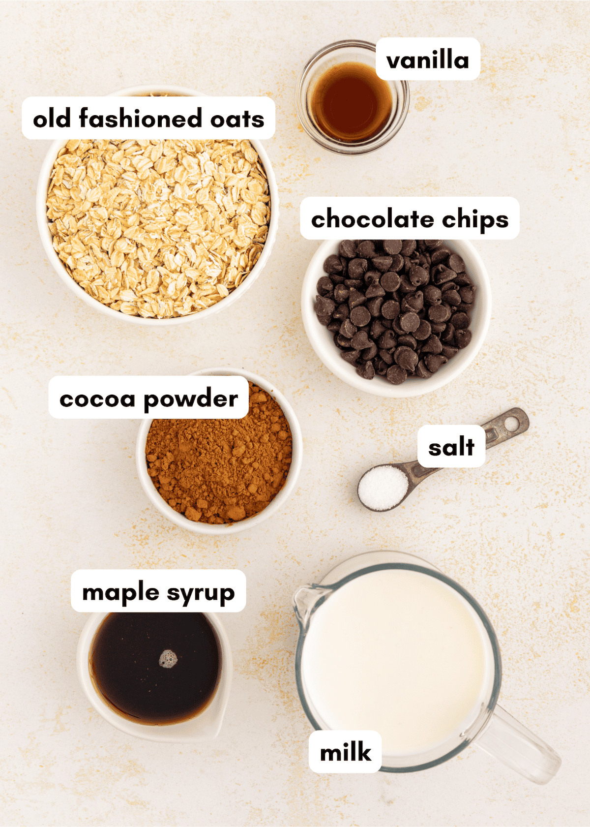 Bowls of chocolate oatmeal, cocoa powder, maple syrup, milk, vanilla, and salt on a table.