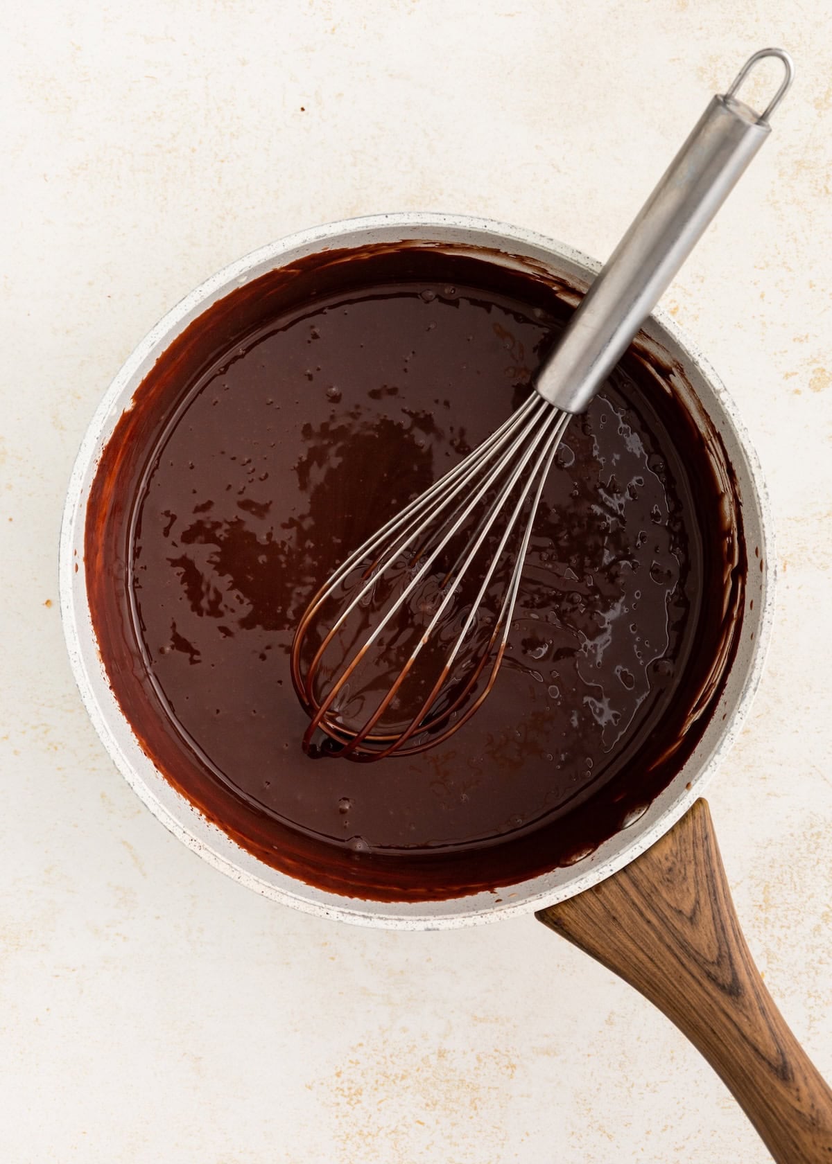 A whisk in a pot of melted chocolate sauce on a light surface.