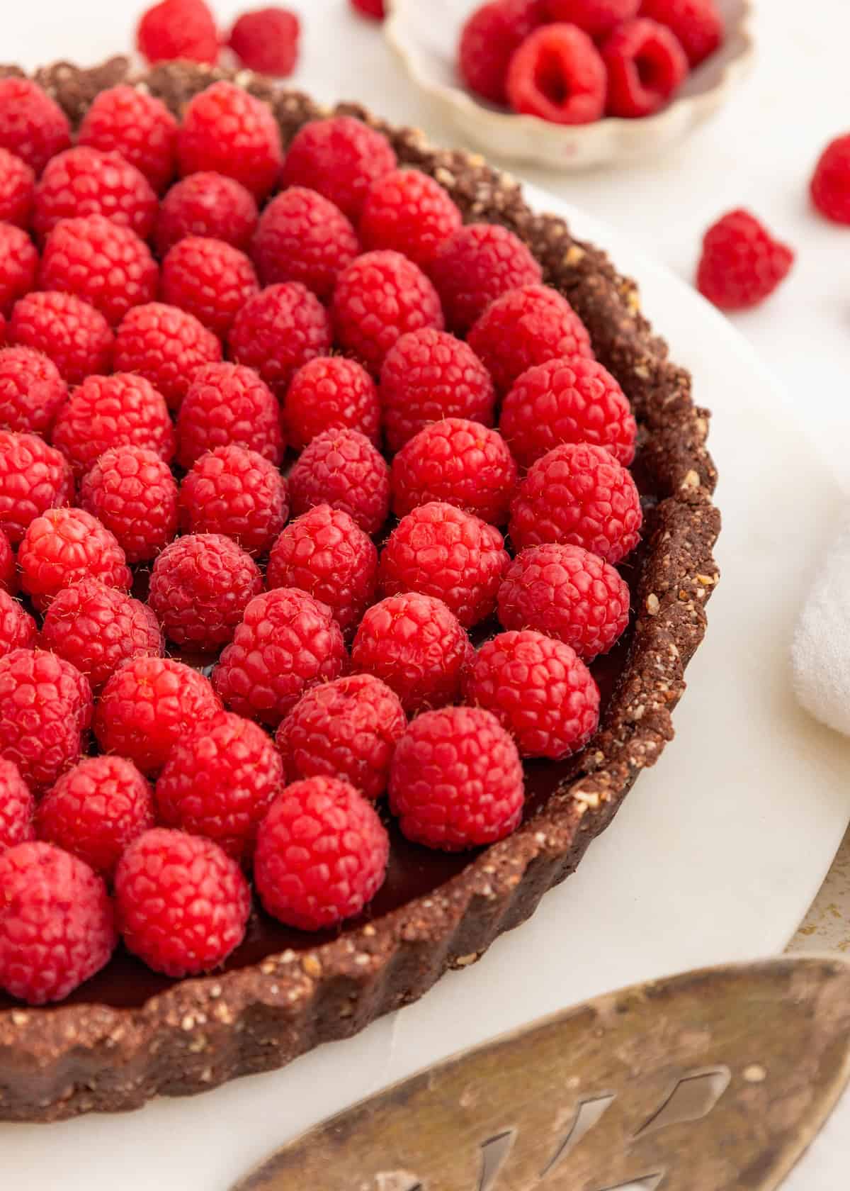 A chocolate tart topped with fresh raspberries on a white surface, with raspberries nearby.