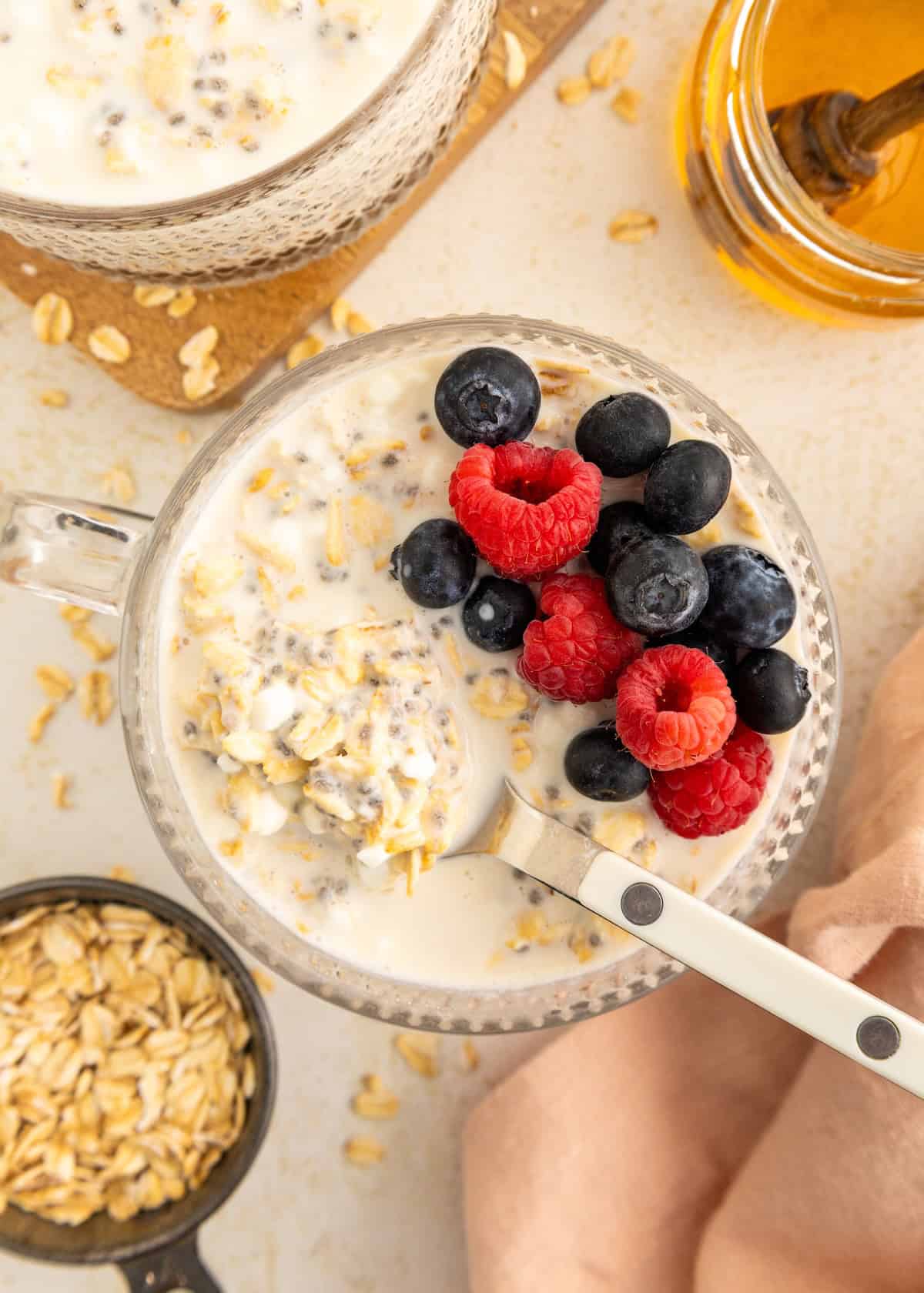 Overnight oats with milk, raspberries, and blueberries in a glass mug, with honey on the side.