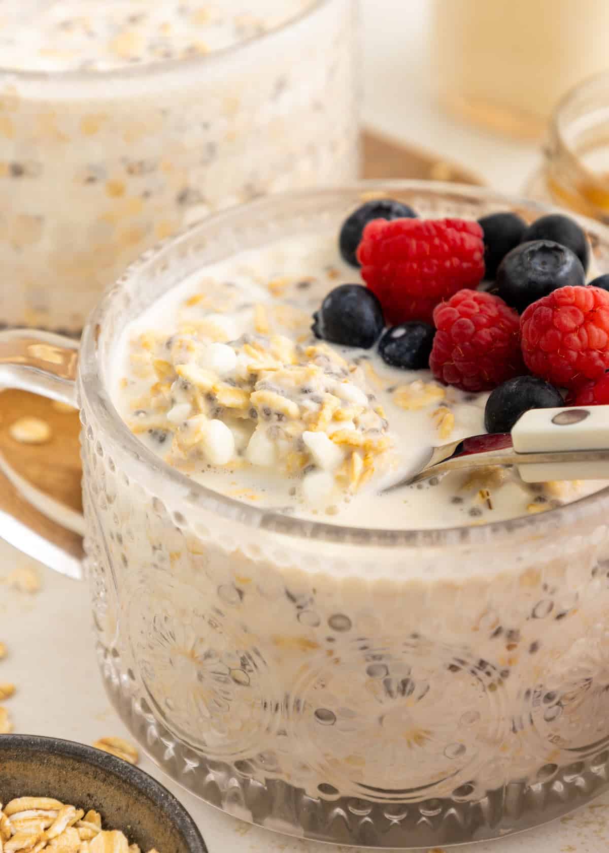 A glass mug of overnight oats topped with blueberries and raspberries with a spoon inside.