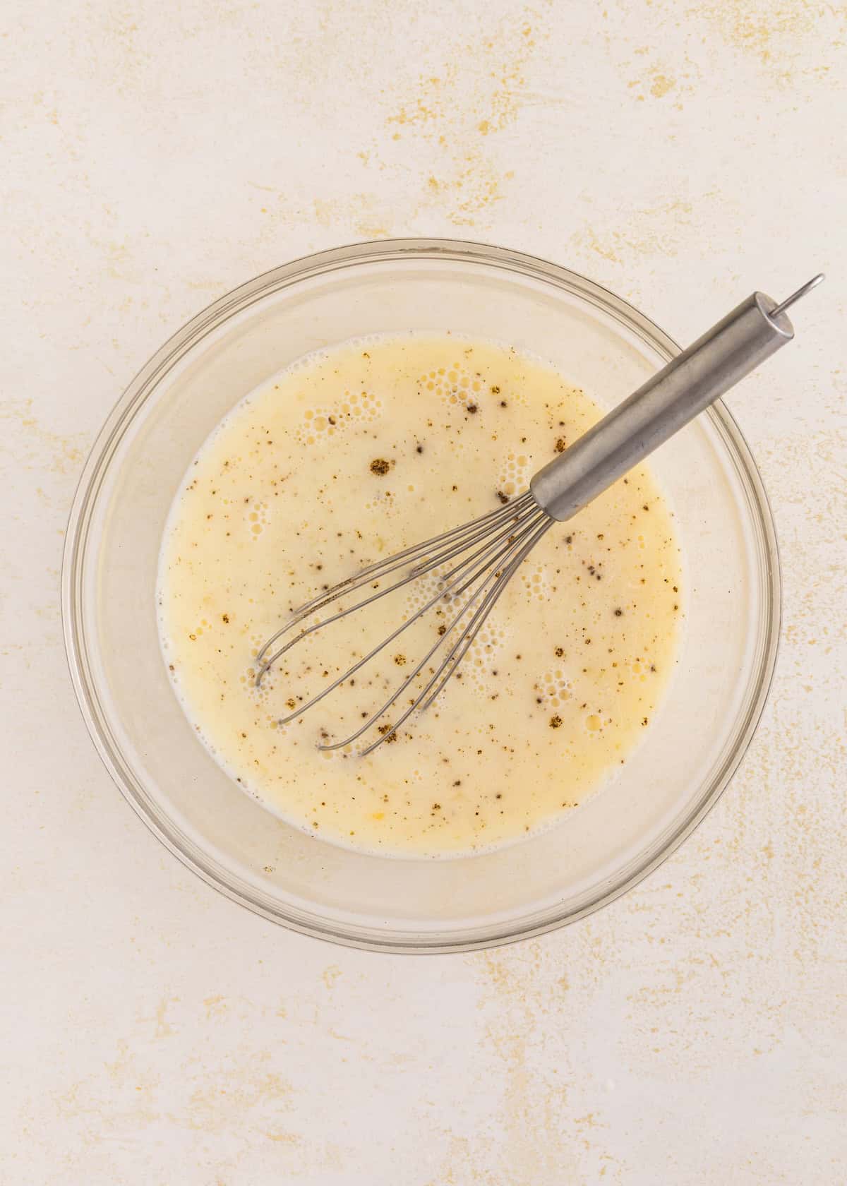 A whisk in a bowl of seasoned beaten eggs on a light surface.