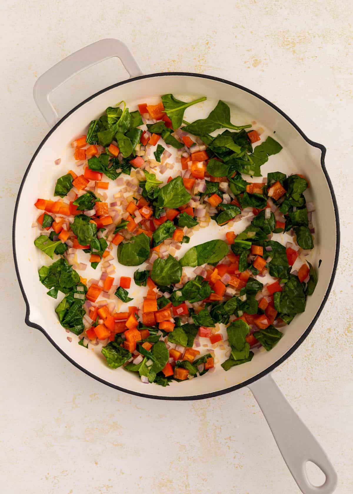 Chopped spinach, tomato, and bell pepper sautéing in a white skillet on a light background.