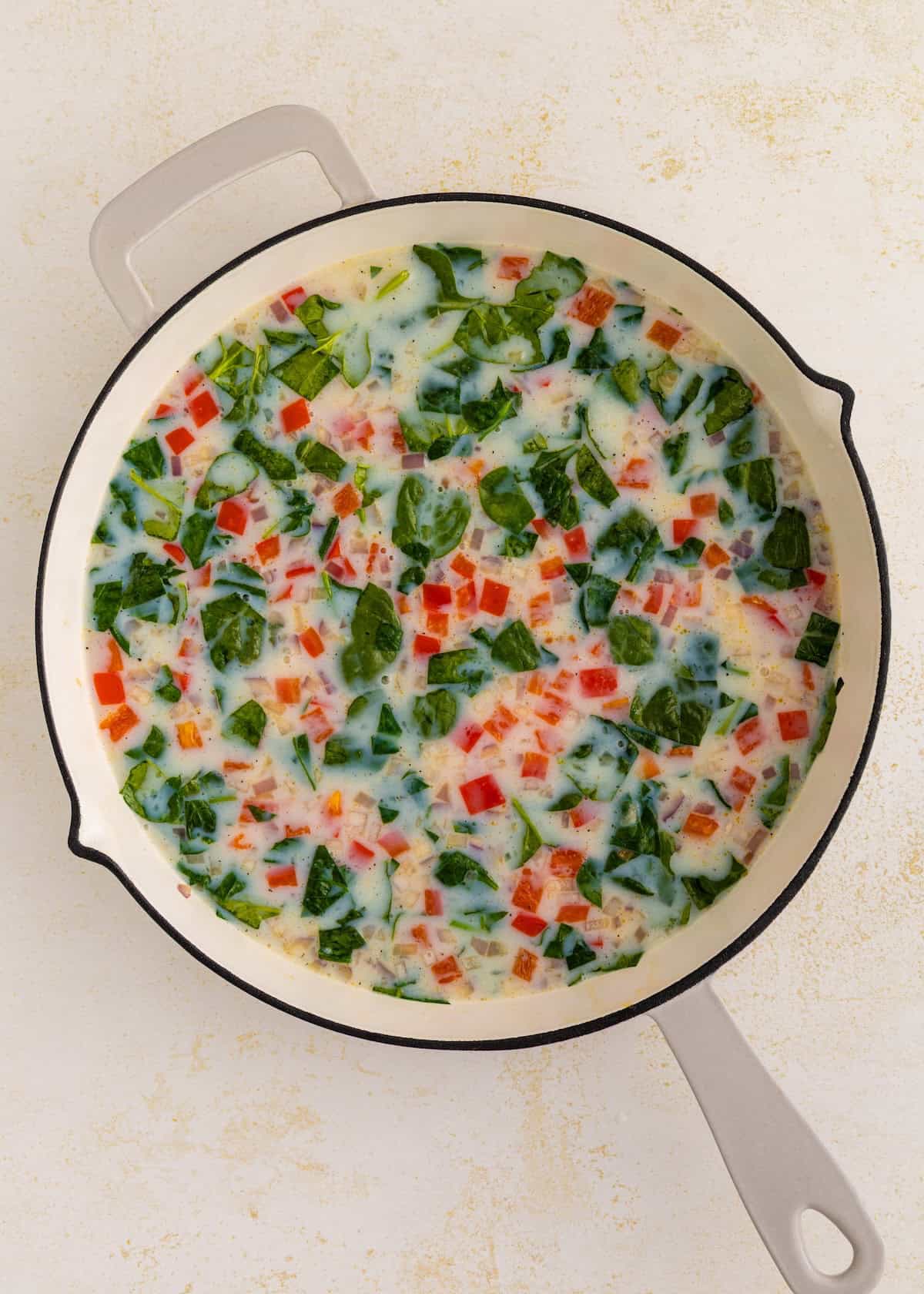 Egg mixture with chopped spinach, red pepper, and onions in a round white skillet.