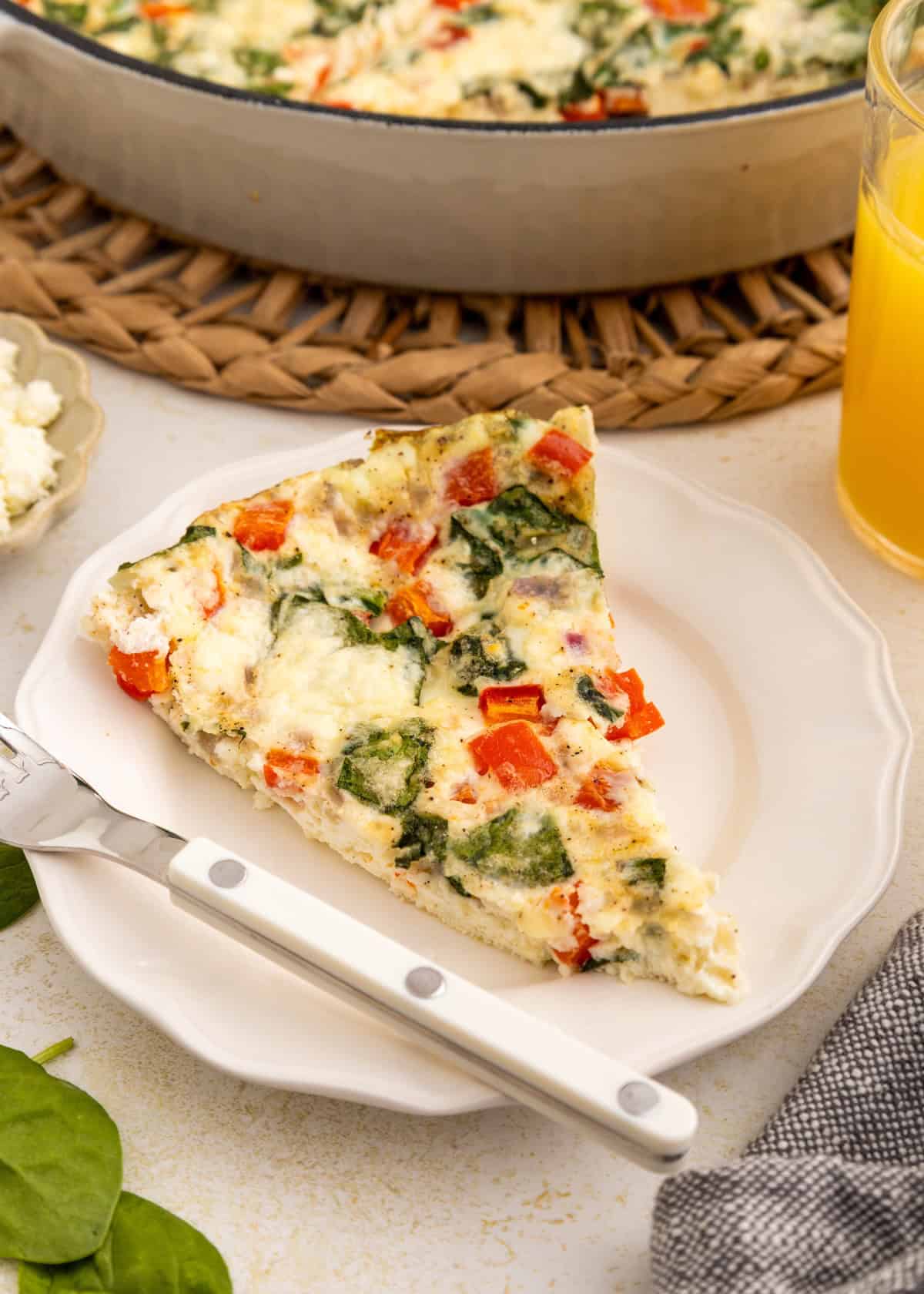 A slice of vegetable frittata on a white plate with a knife and a glass of orange juice.