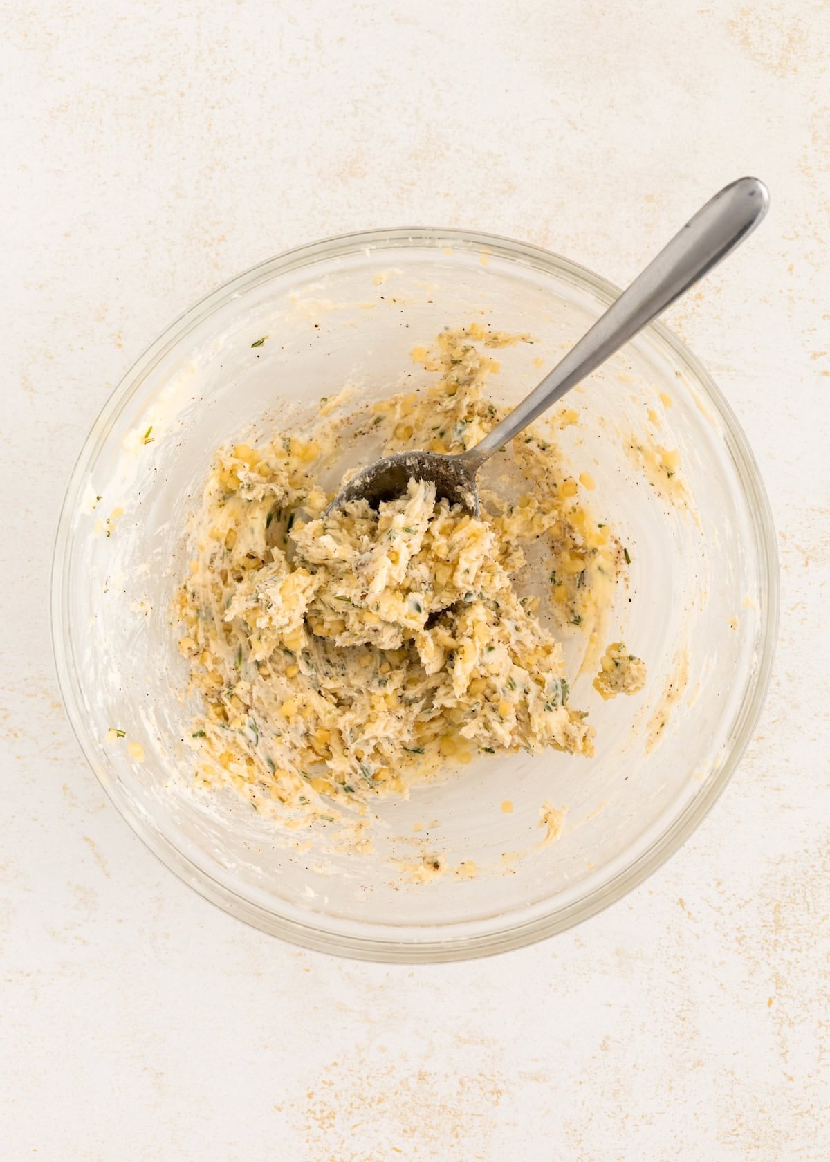 A glass bowl with garlic herb butter mixture for roast beef, with a spoon on a light surface.
