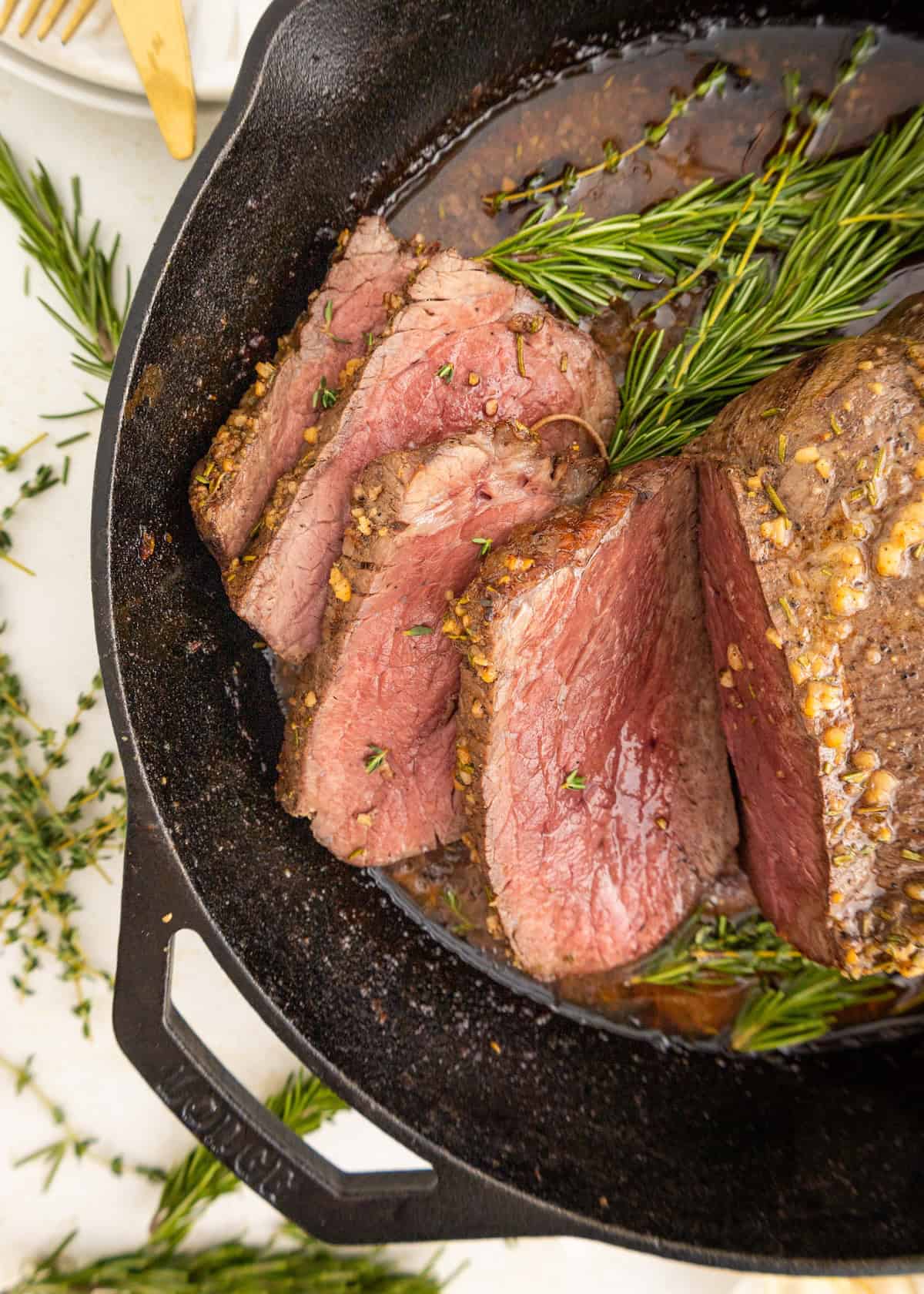 Sliced garlic herb roast beef steak with rosemary and thyme in a cast iron skillet.