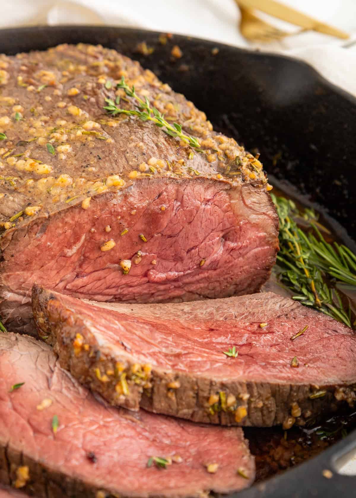Garlic herb roast beef sliced in a skillet, garnished with fresh herbs and garlic.