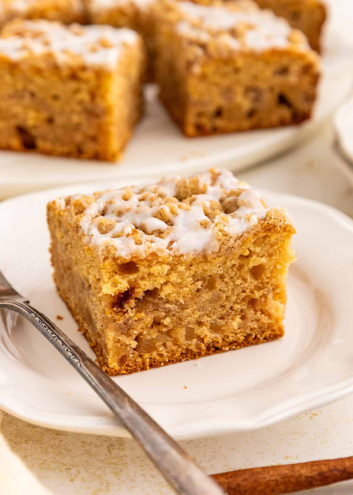 A slice of gluten-free coffee cake on a white plate with a fork, more cake in the background.