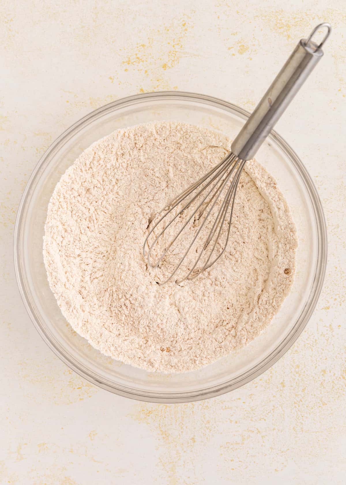 A glass bowl of gluten-free pumpkin cake flour mixture with a metal whisk on a light surface.