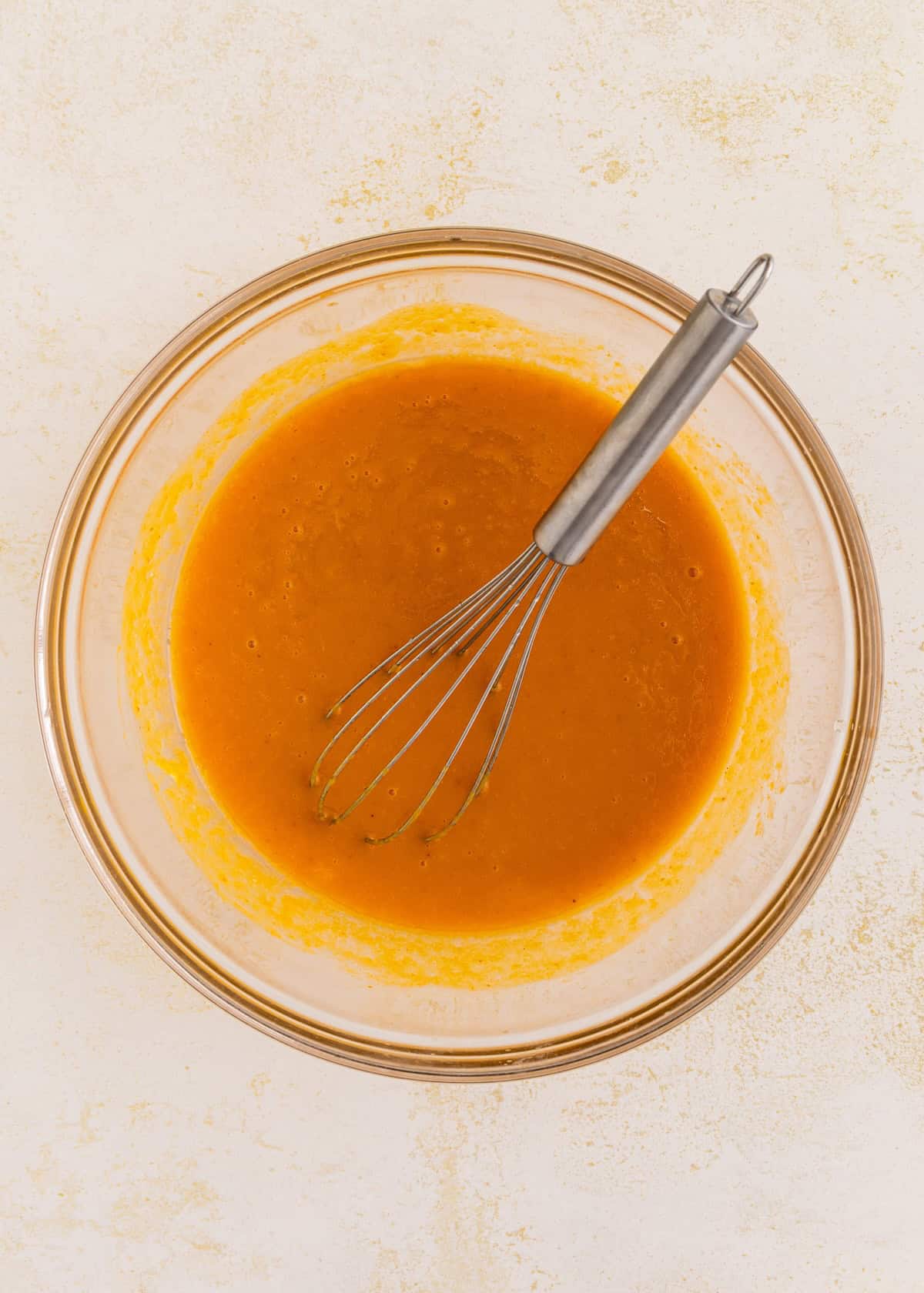 Glass bowl with orange mixture for gluten-free pumpkin cake and metal whisk on light surface.