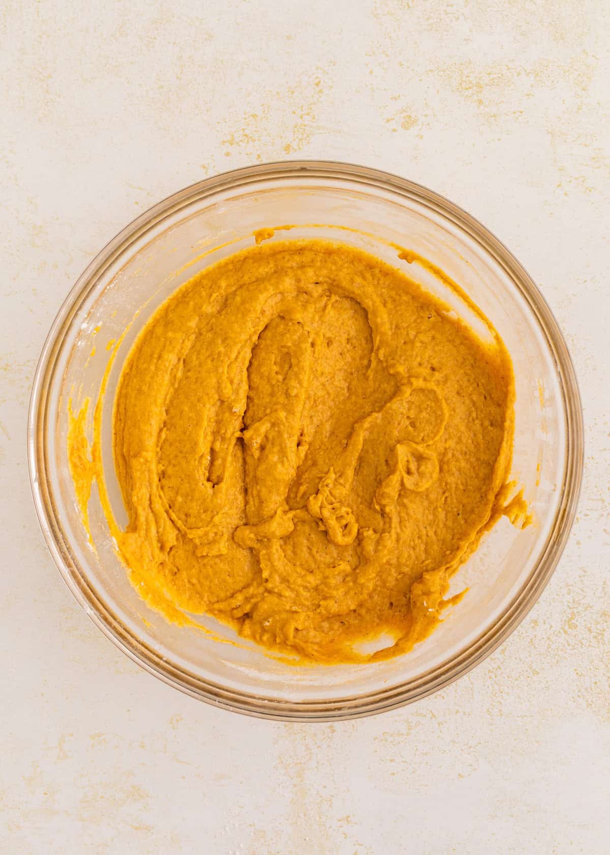 A glass bowl filled with smooth, orange gluten-free pumpkin cake batter on a light surface.