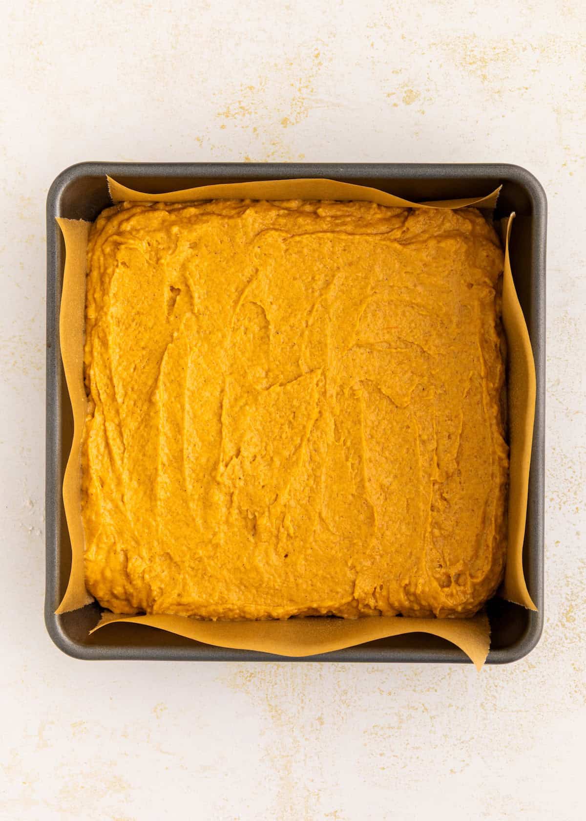 Square baking pan with parchment, filled with gluten-free pumpkin cake batter on a white surface.