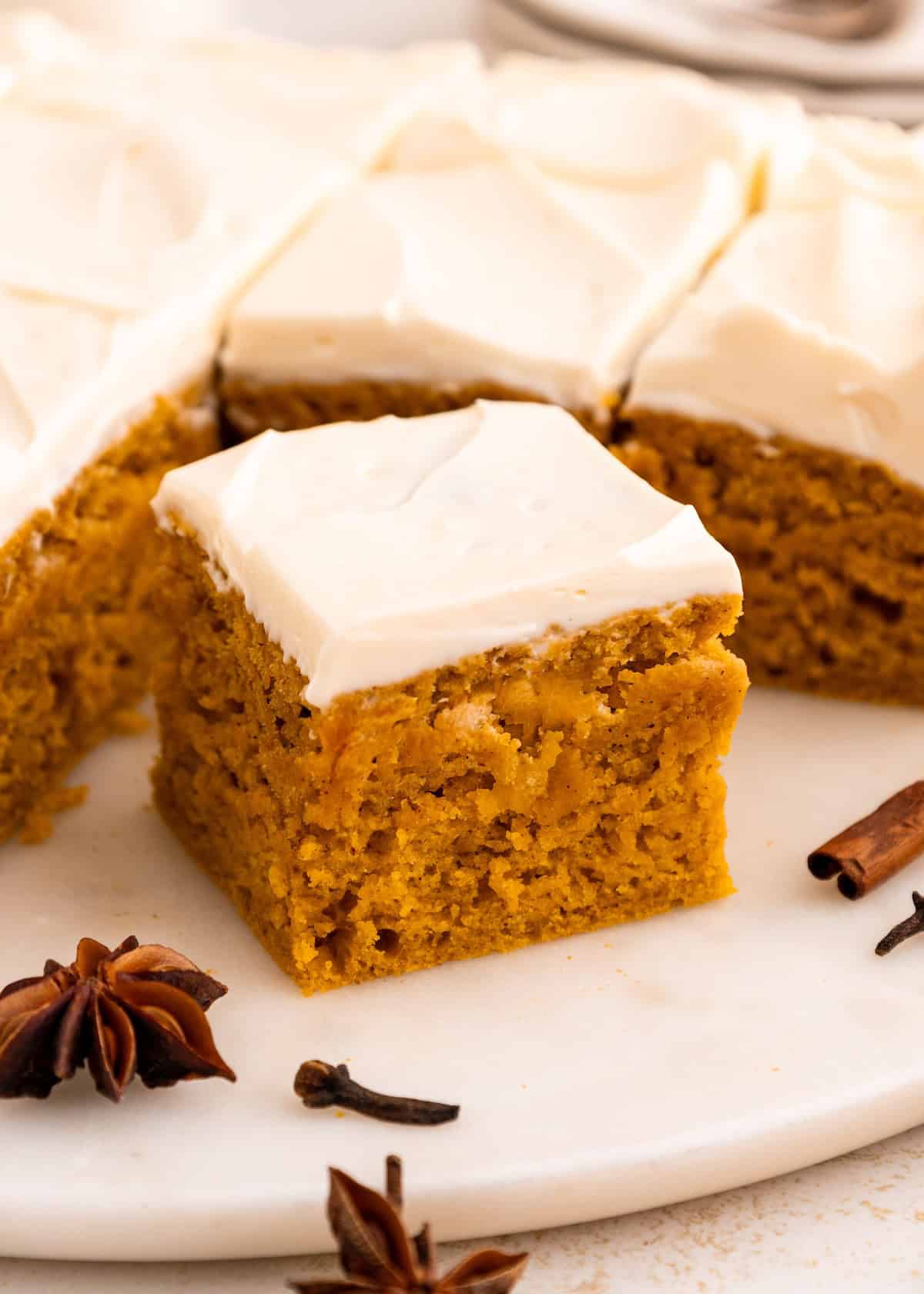A slice of gluten-free pumpkin cake with white frosting, on marble and surrounded by spices.