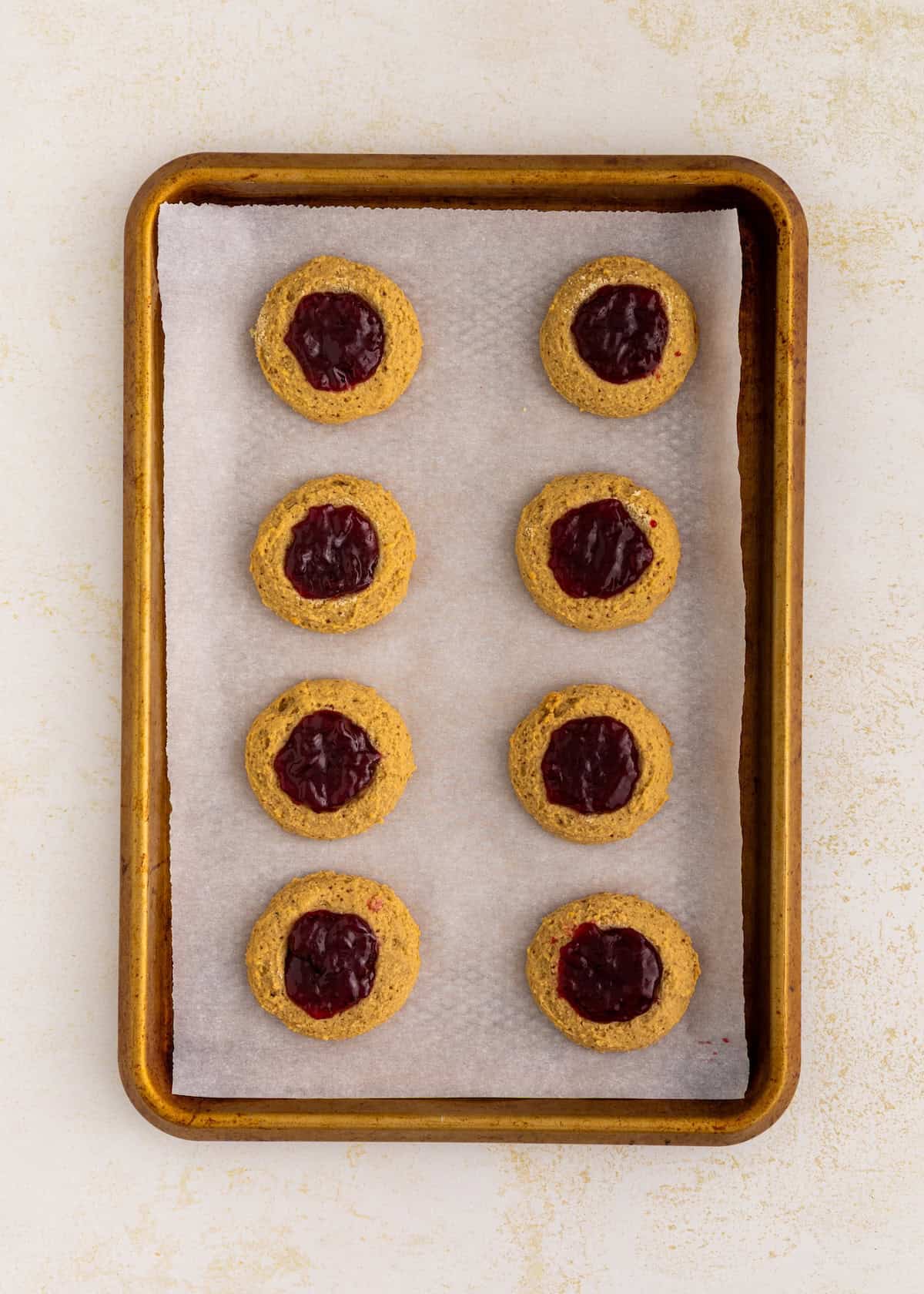 A baking tray with eight gluten-free thumbprint cookies filled with red jam on parchment paper.
