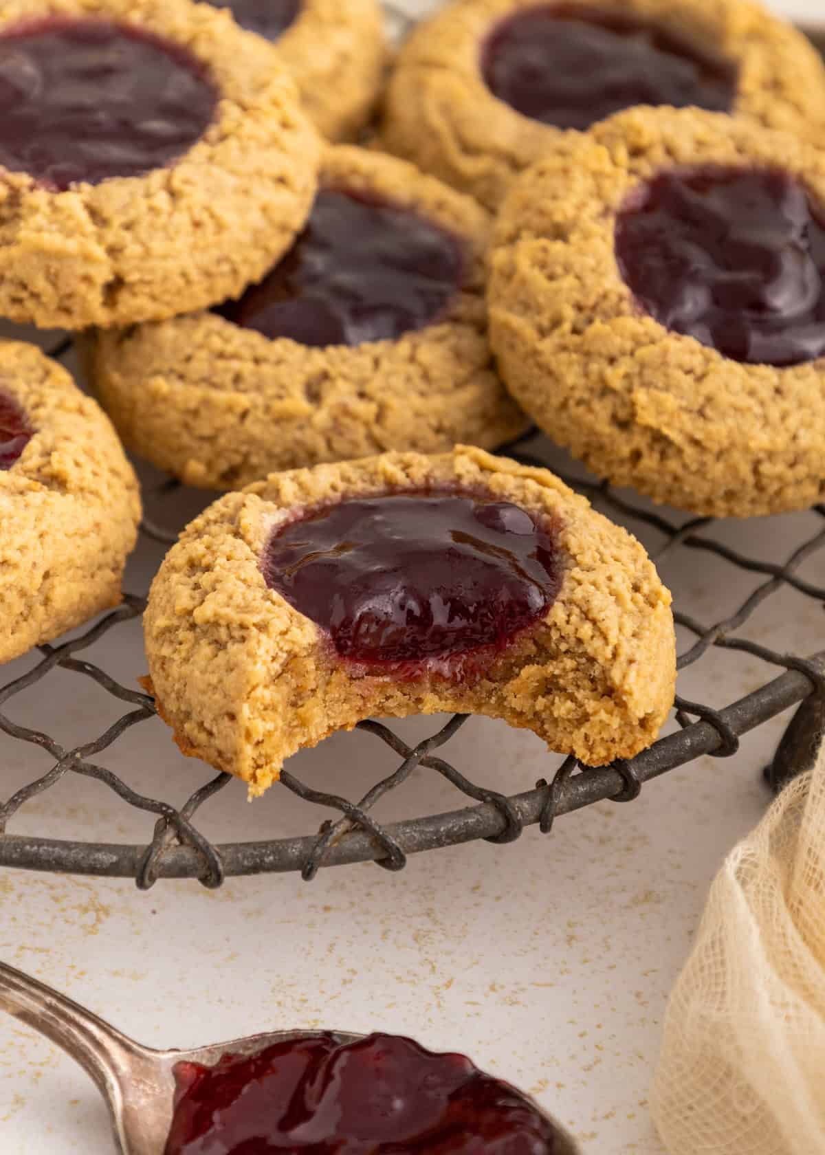 A bitten gluten-free thumbprint cookie with jam cools on a rack among other cookies.