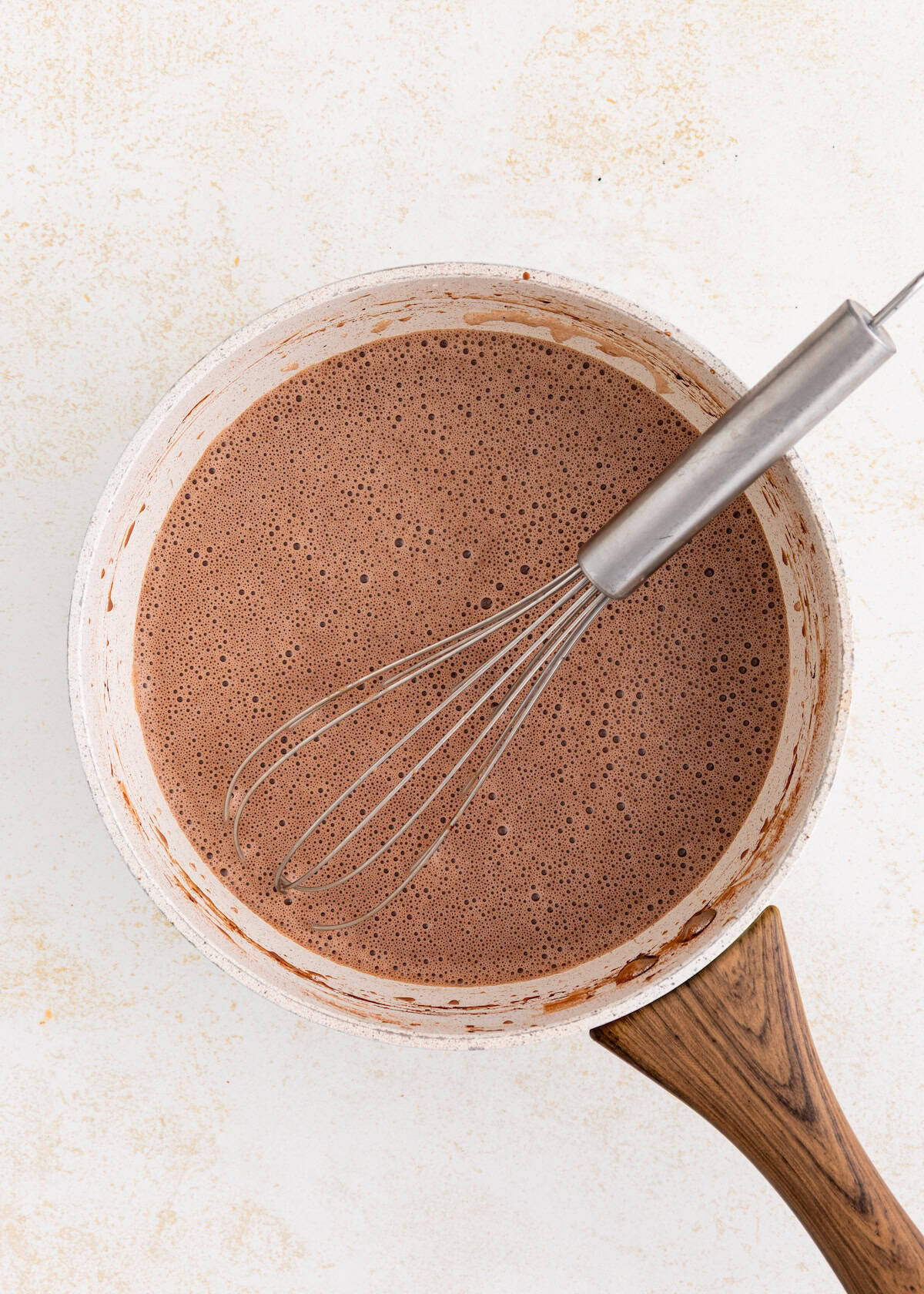 A whisk in a pot of frothy homemade fudgesicles mixture on a light surface.