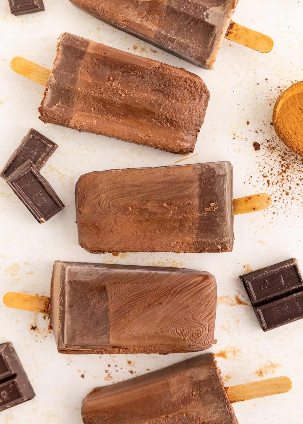 Four homemade fudgesicles on a white surface with chocolate pieces and cocoa powder scattered around.