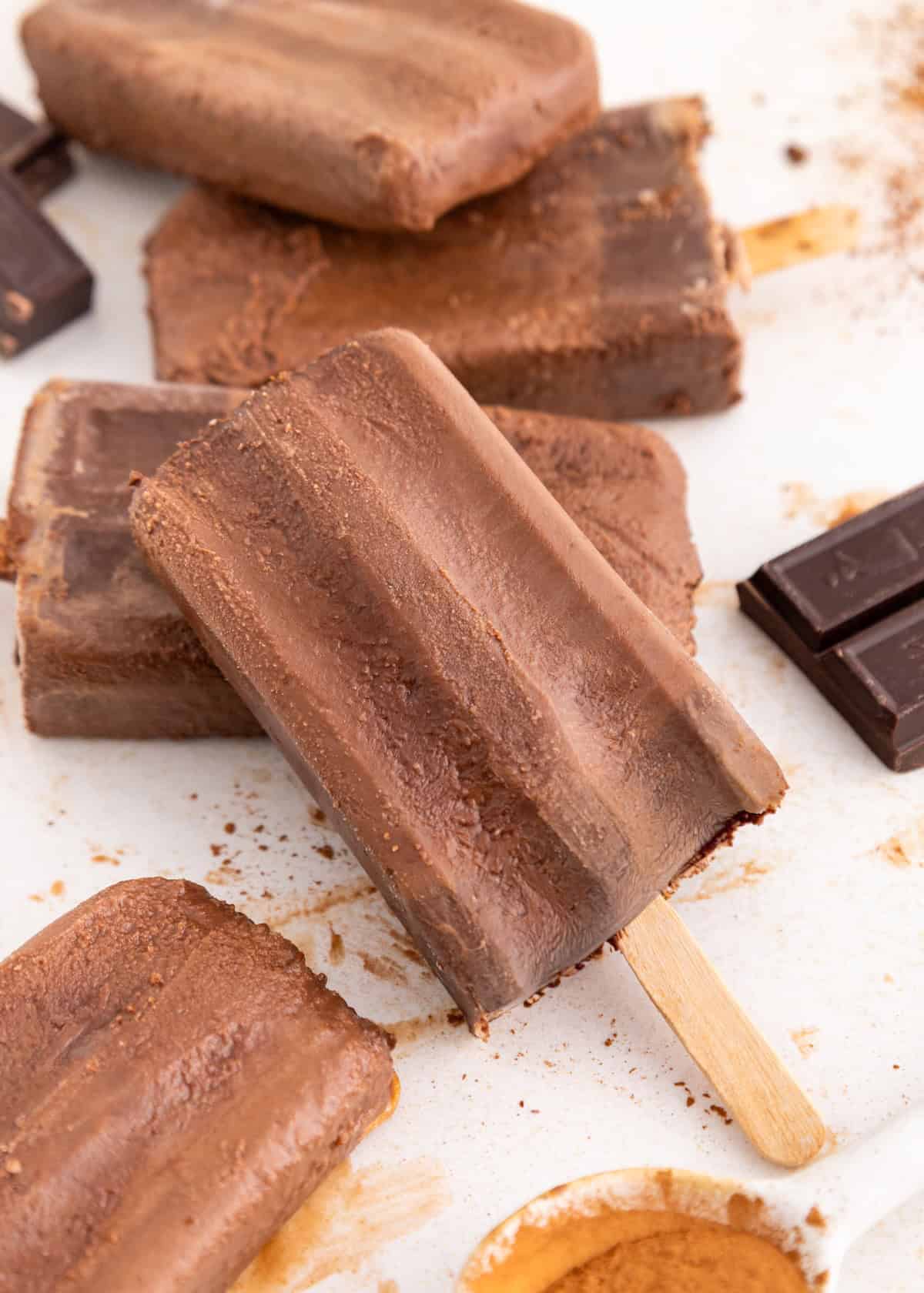 Homemade fudgesicles on a white surface with chocolate pieces and cocoa powder nearby.