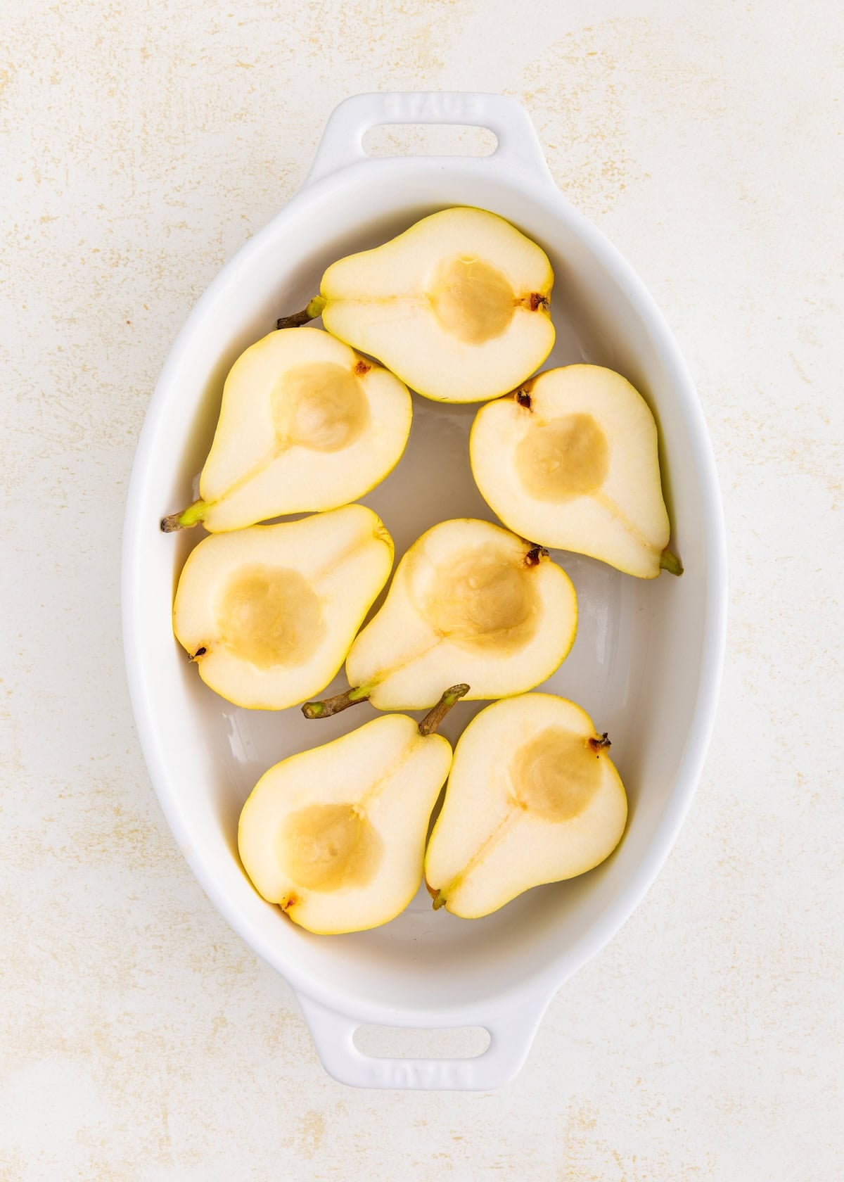 Eight honey baked pears arranged in a white oval baking dish on a light background.