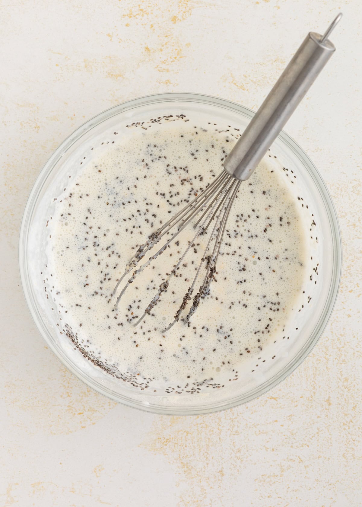 A glass bowl with Protein Chia Pudding mixture and a metal whisk on a light surface.