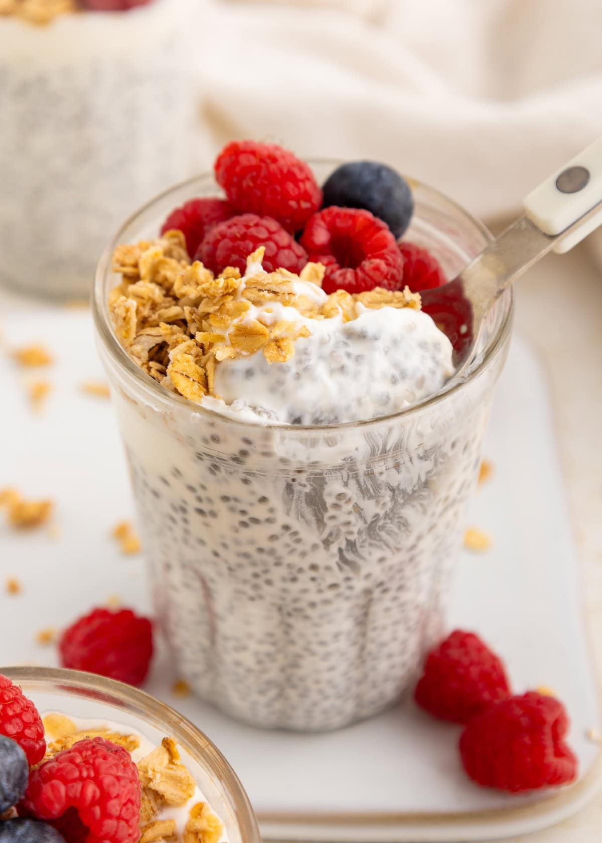 A glass of Protein Chia Pudding topped with granola, raspberries, and blueberries with a spoon.