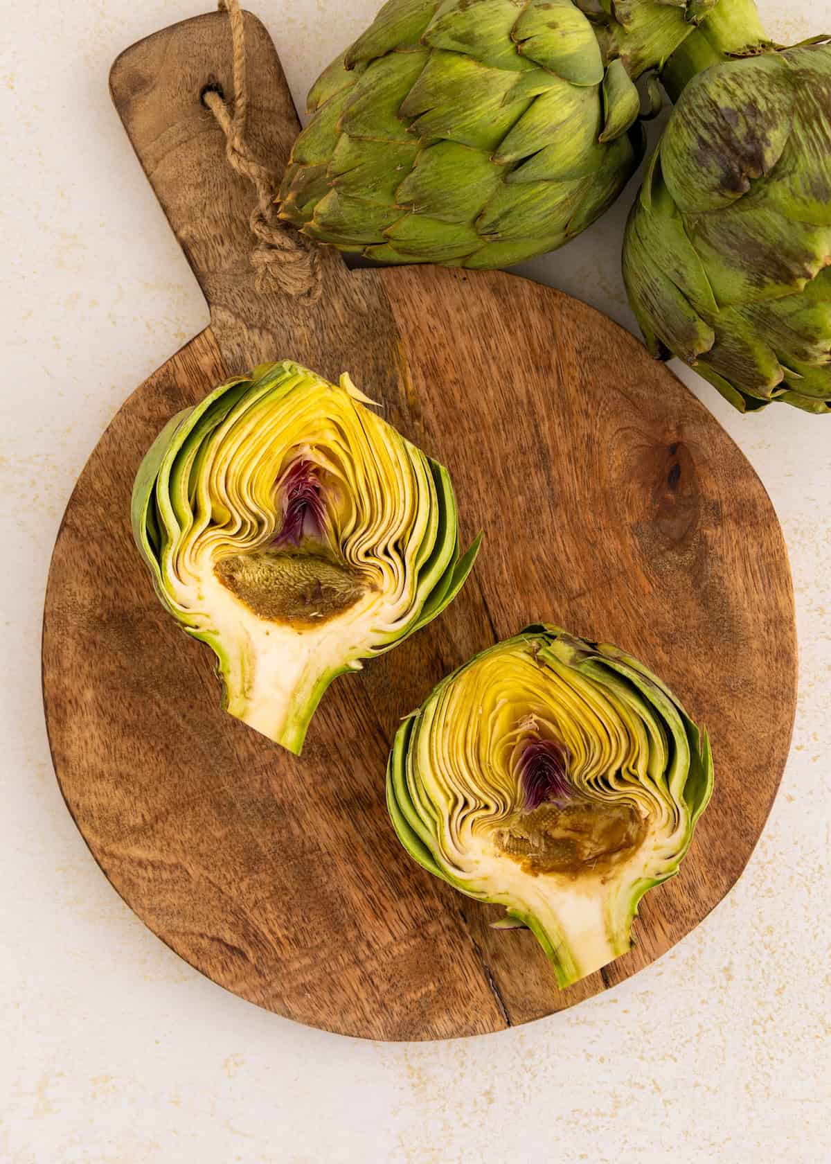 Two artichoke halves on a wooden cutting board, whole artichokes beside them.