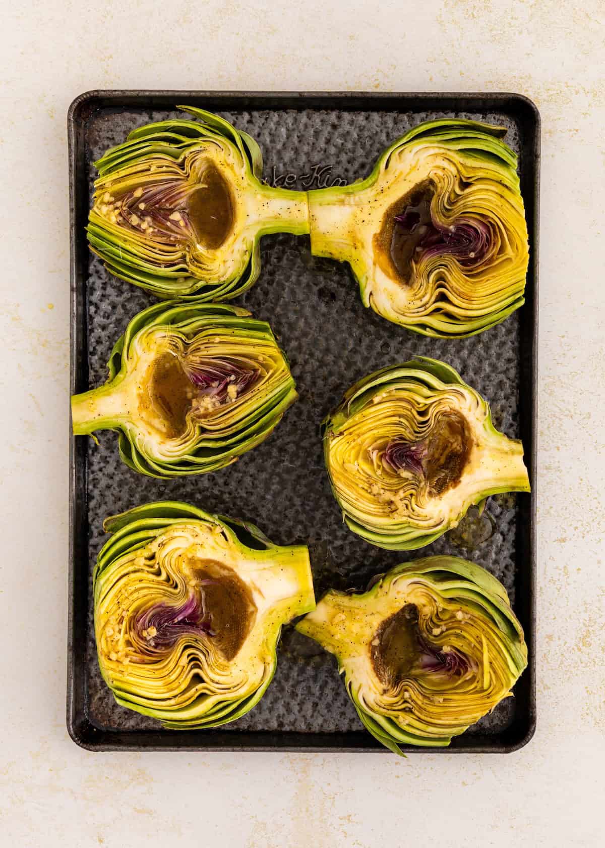 Five halved artichokes on a baking tray, seasoned and ready for roasting.