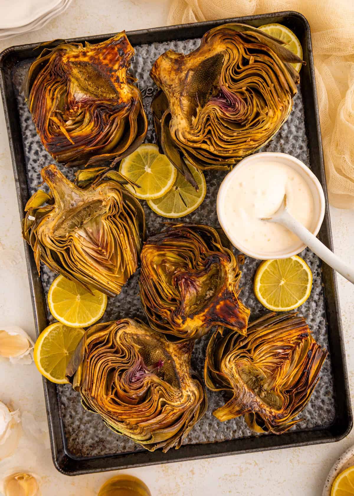 Roasted artichoke halves with lemon slices and a bowl of dipping sauce on a metal tray.