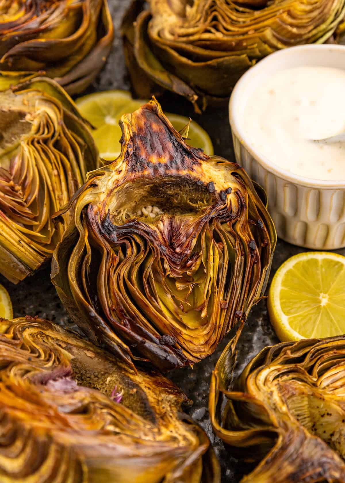 Grilled artichoke halves with lemon slices and a small bowl of creamy dipping sauce.