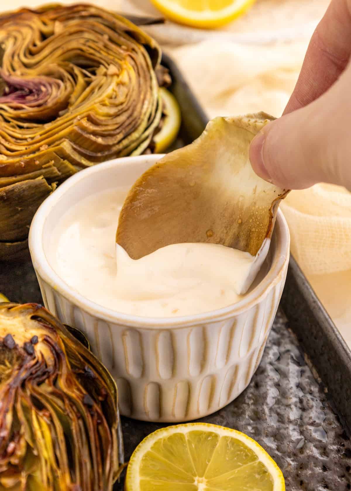 A hand dips an artichoke leaf into a bowl of creamy white sauce, with lemon slices nearby.