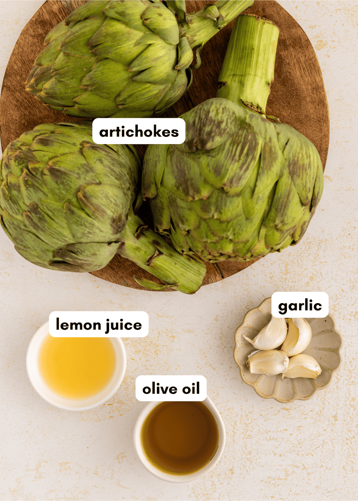 Artichokes, lemon juice, olive oil, and garlic displayed on a light surface.