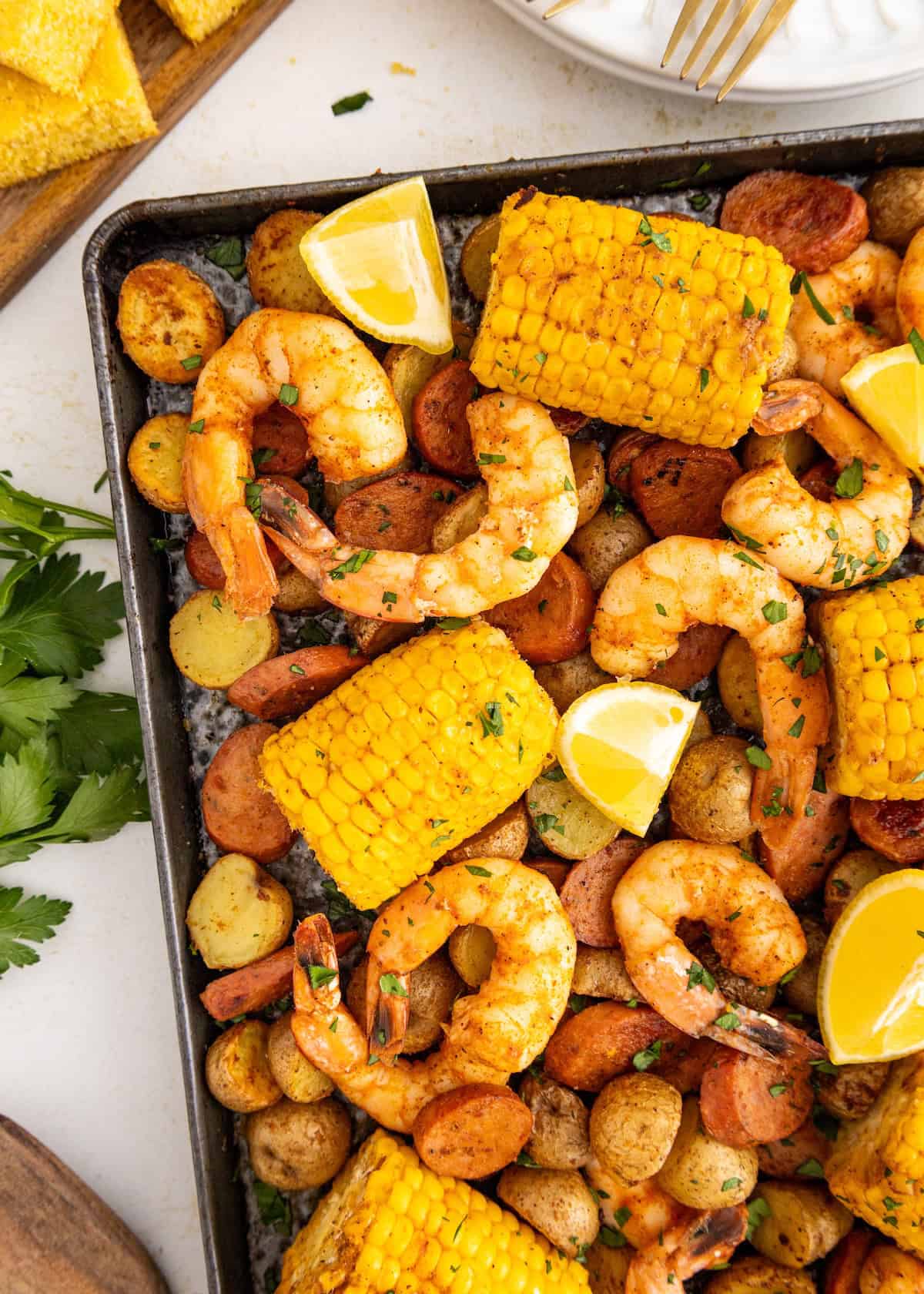 Sheet pan with shrimp, corn, sausage, potatoes, and lemon wedges garnished with herbs.
