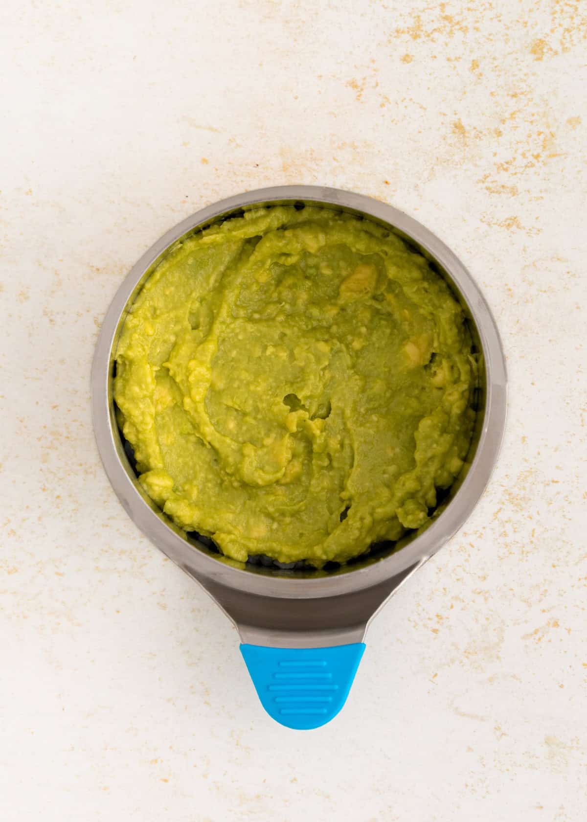 A metal bowl with mashed guacamole for sushi stacks, featuring a blue handle on a light countertop.