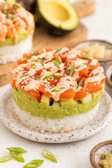 Stacked sushi stacks with rice, avocado, and salmon, topped with sauce, sesame seeds, and green onions.